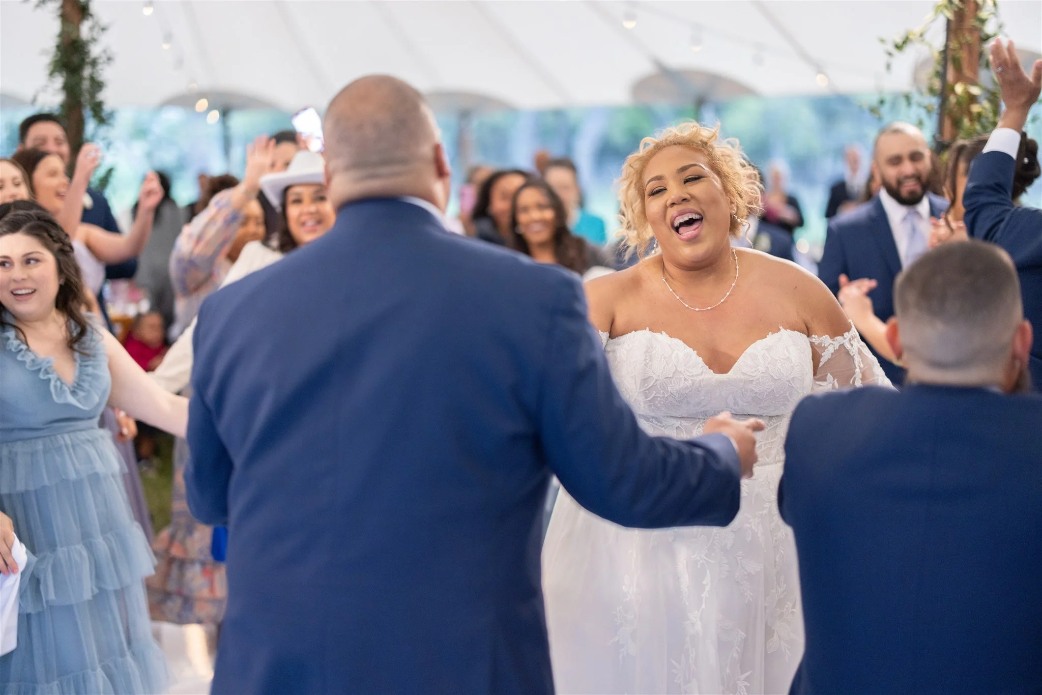 A bride dancing with her groom at her wedding reception, surrounded by smiling guests in a decorated outdoor tent.