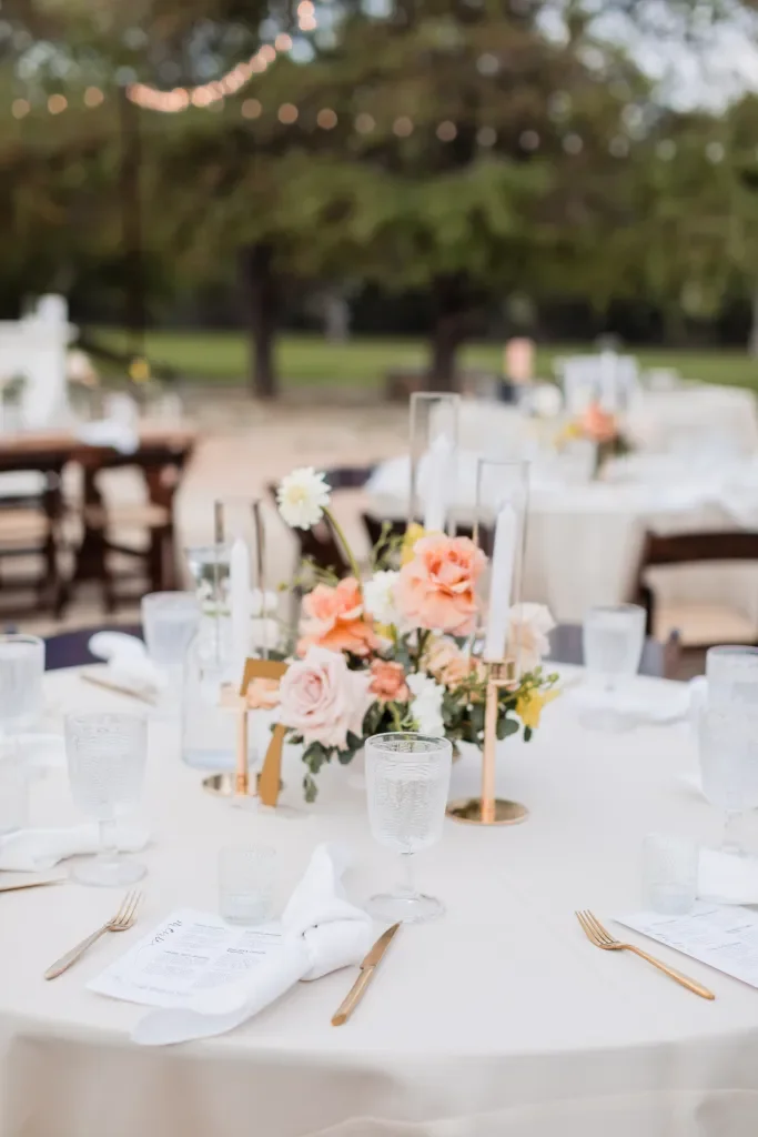 Outdoor wedding reception table decorated with pink and white flowers, candles, and glassware, set in a park with string lights overhead.