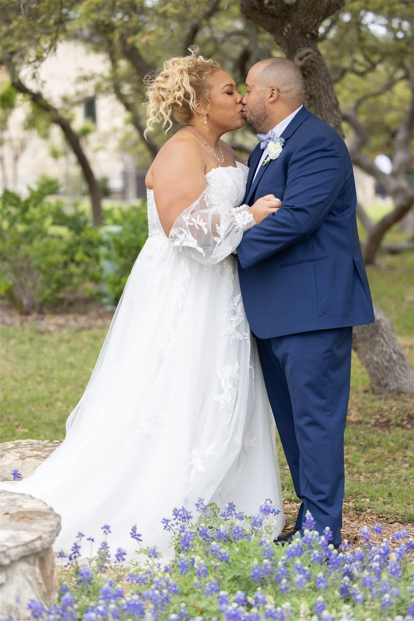 A bride and groom share a kiss outdoors, with the bride in a white wedding dress and the groom in a navy suit, surrounded by green trees and purple flowers.