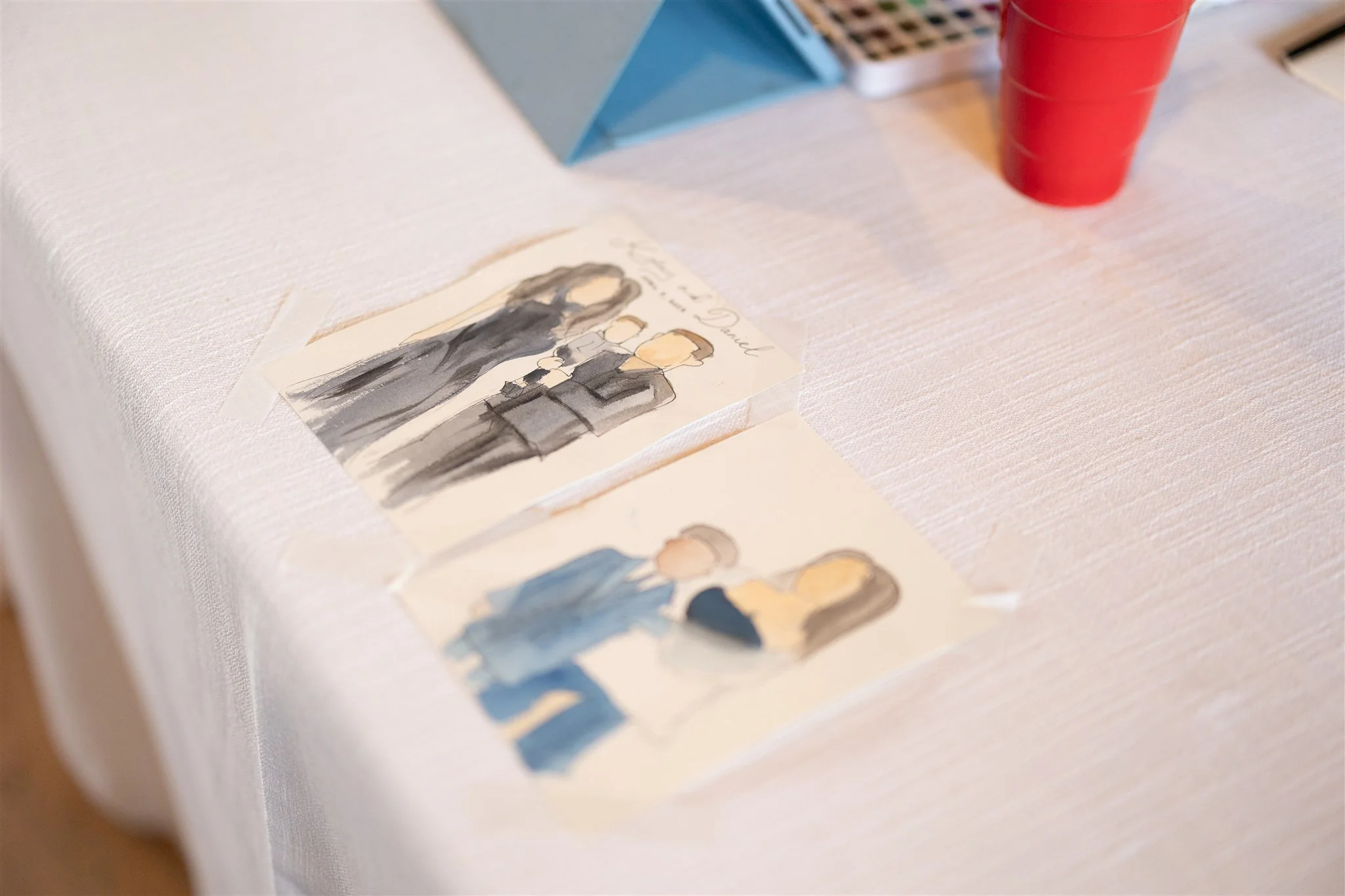 Two watercolor paintings of couples, taped to a white tablecloth, with red plastic cups and a blue geometric box in the background.