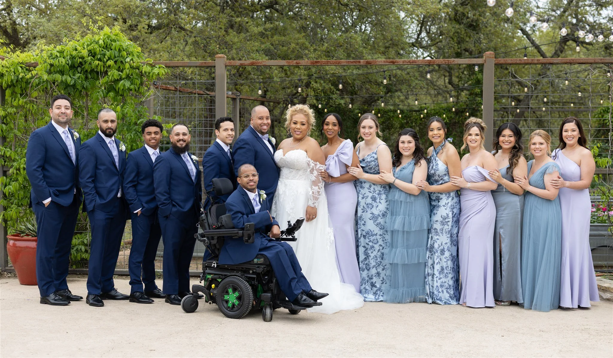 A diverse group of people dressed in formal attire standing outdoors for a wedding photo, a bride in a white wedding dress and a groom in a blue suit at the center, with bridesmaids and groomsmen standing on either side, one man in a wheelchair in fr