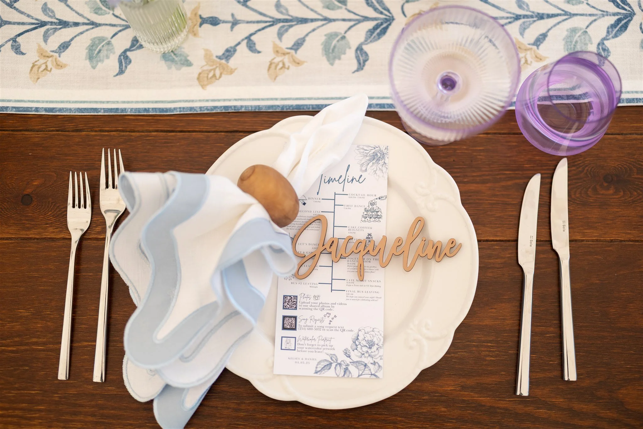 A decorated table setting with a white plate, utensils, and purple glasses, featuring a party or wedding theme with a name card reading 'Jacqueline' and a menu, on a wooden table with a blue and white floral table runner.