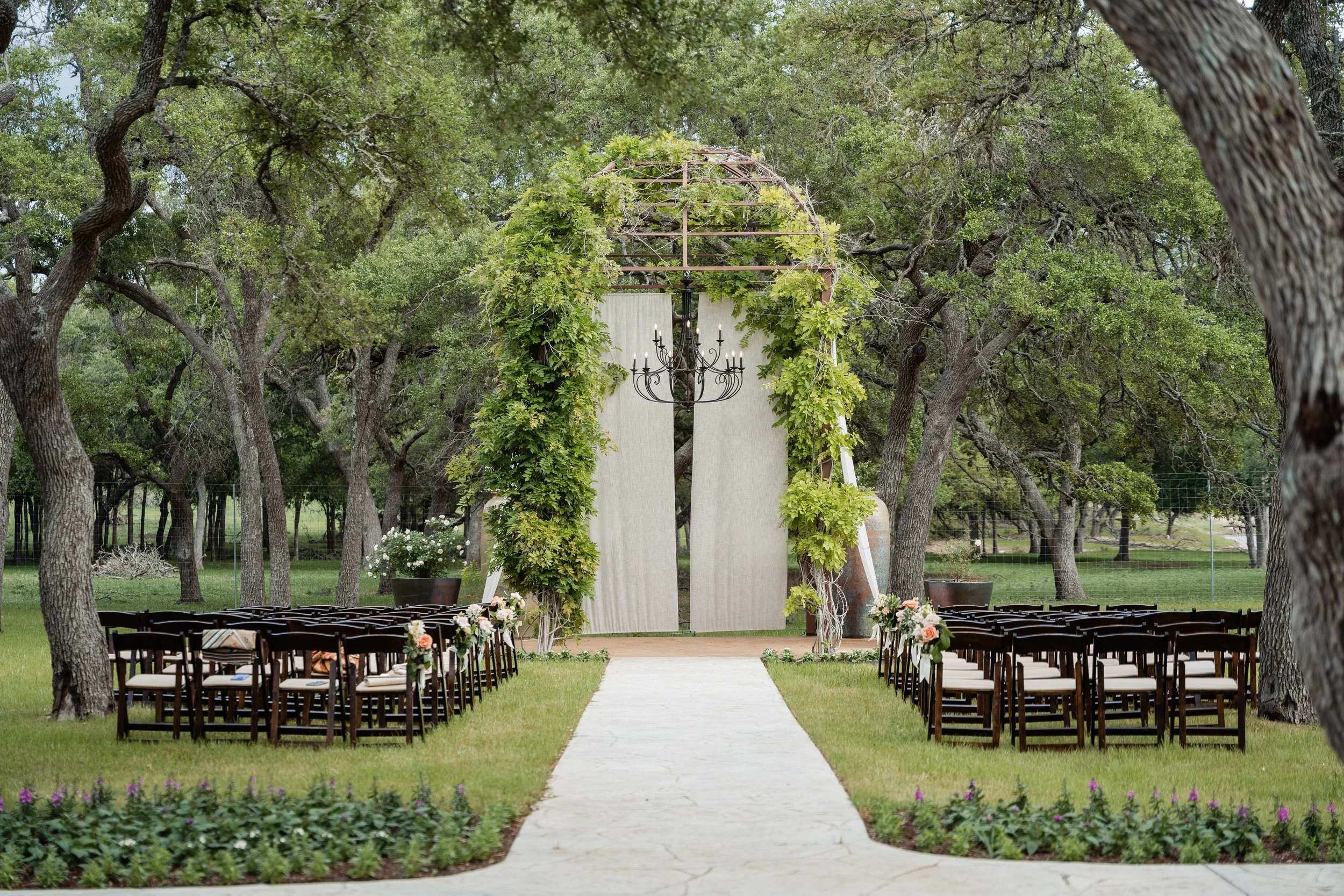 Outdoor wedding ceremony setup with chairs, flower decorations, a white aisle, and a floral arch under trees.