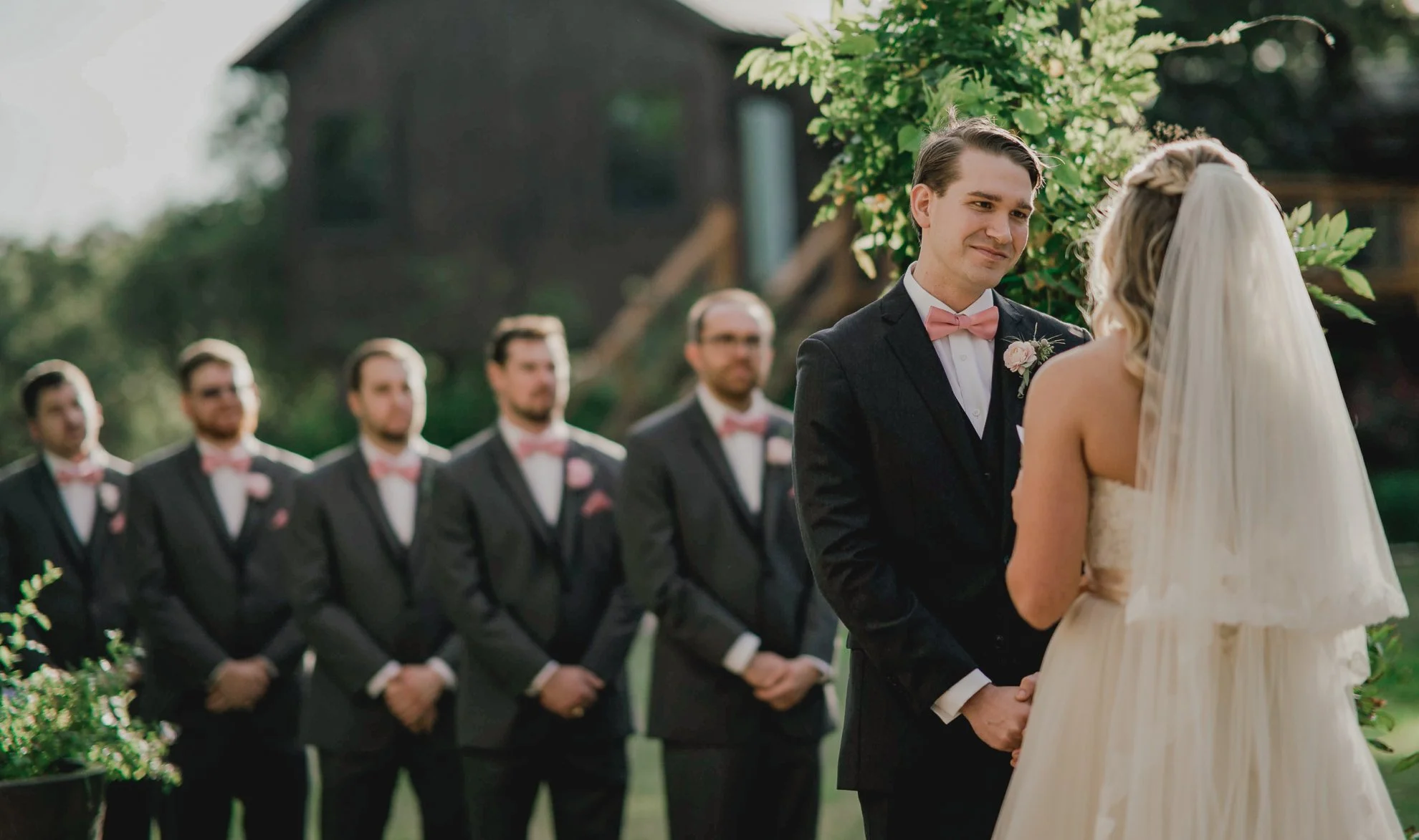A bride and groom exchanging vows outdoors, with the groom holding the bride's hands, in front of bearded men in suits with pink bow ties and boutonnieres, standing in a line.