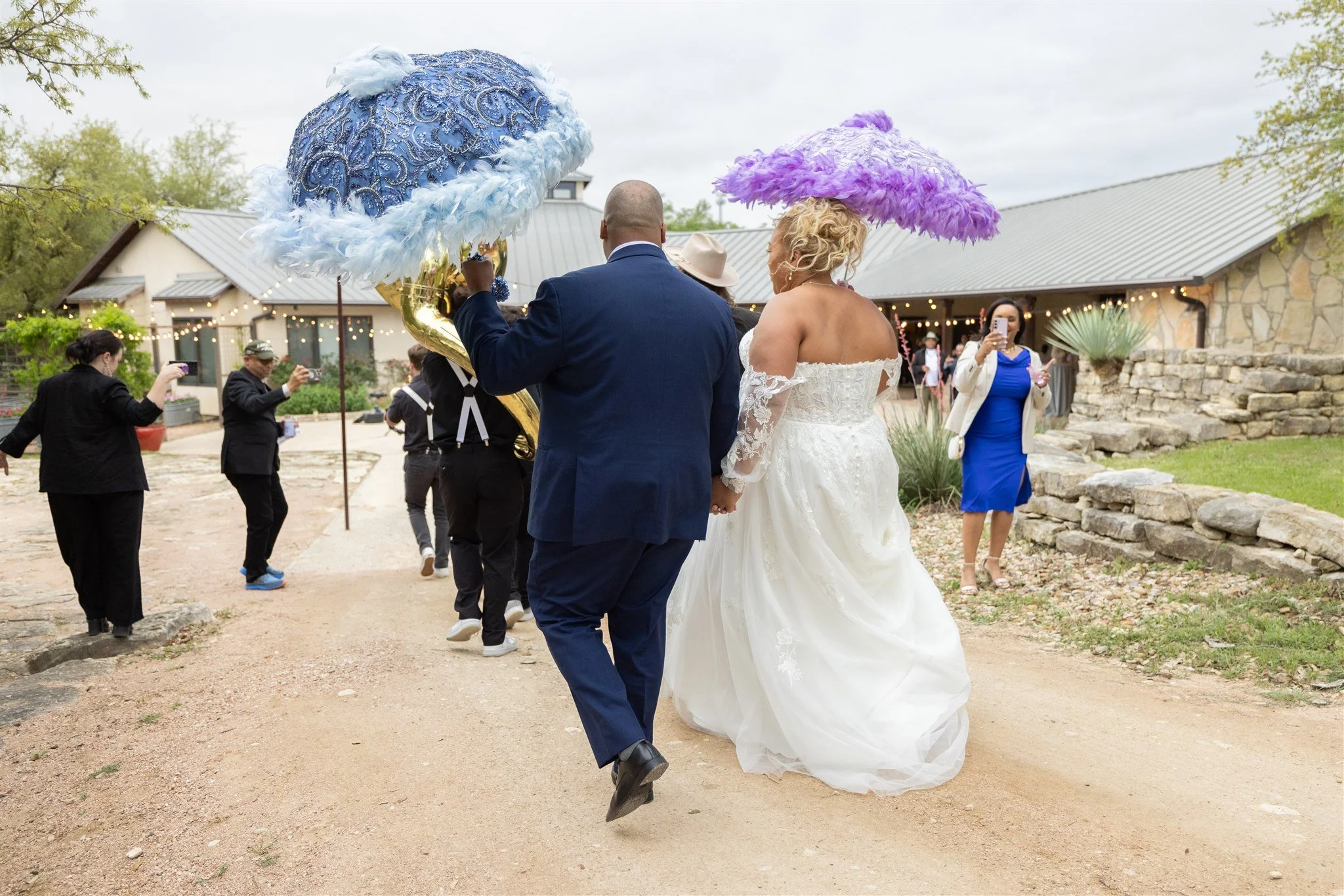 Wedding couple holding hands and walking outdoors on a dirt path, surrounded by guests, some holding umbrellas, in front of a building with string lights, cloudy sky, and greenery.