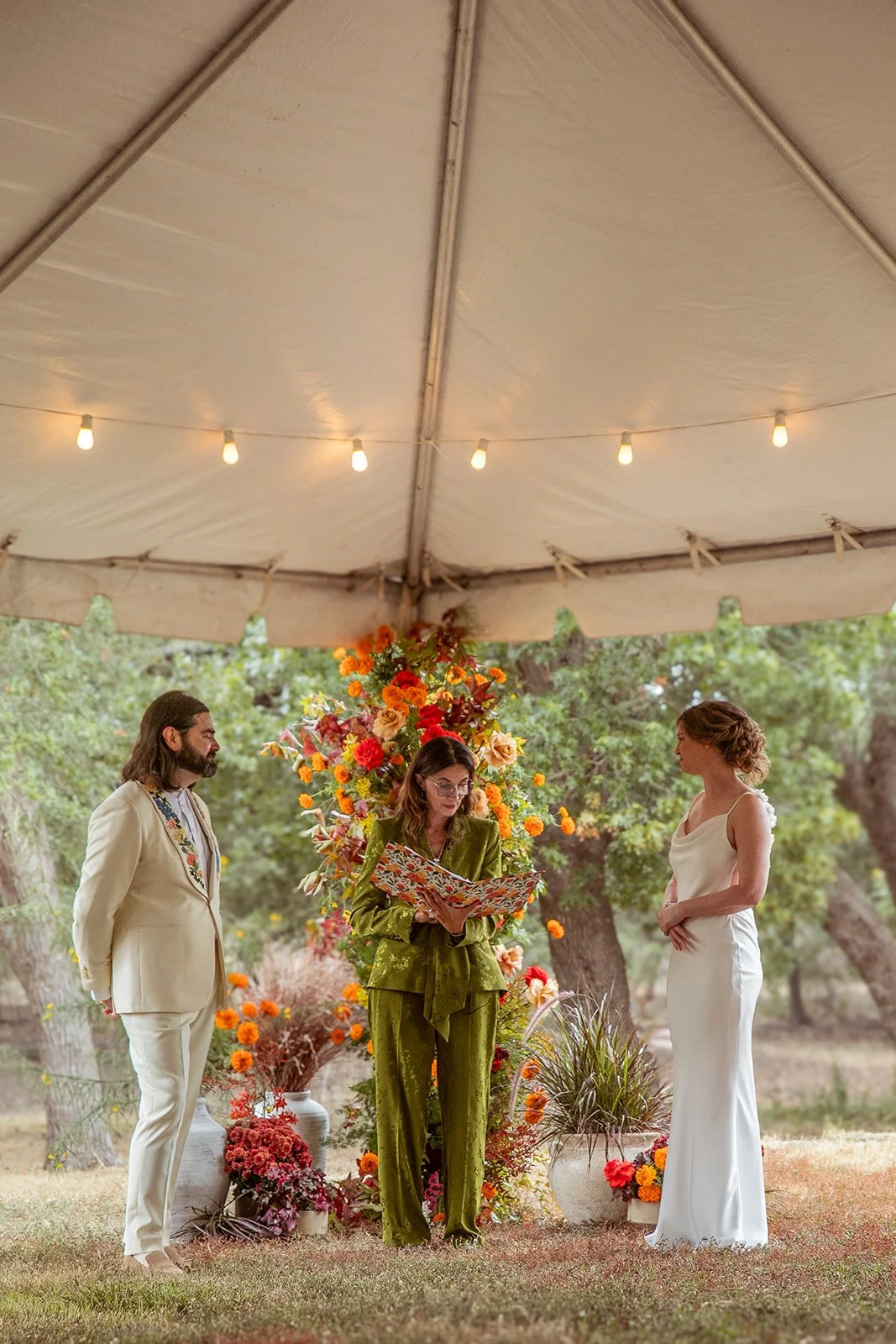 A couple at their wedding ceremony standing on grass under a tent with string lights, with an officiant reading from a book. The scene is decorated with vibrant orange and yellow flowers, potted plants, and trees in the background.