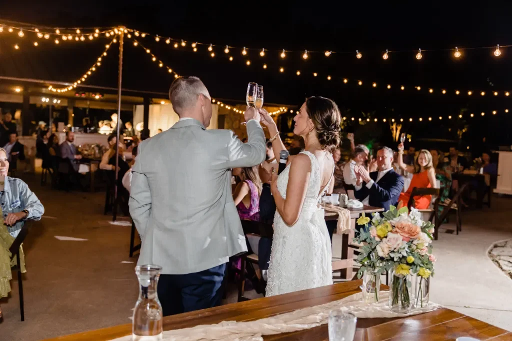 Couple toasting with champagne glasses at outdoor wedding reception under string lights at night
