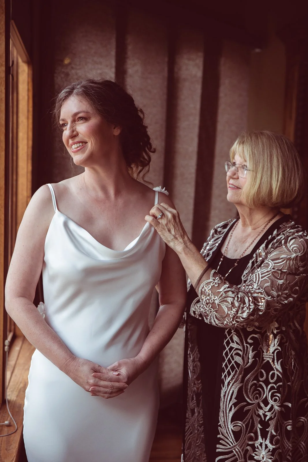 A woman in a white dress is getting ready for a special occasion, assisted by an older woman wearing glasses and a detailed black and silver embroidered jacket, both smiling near a window.