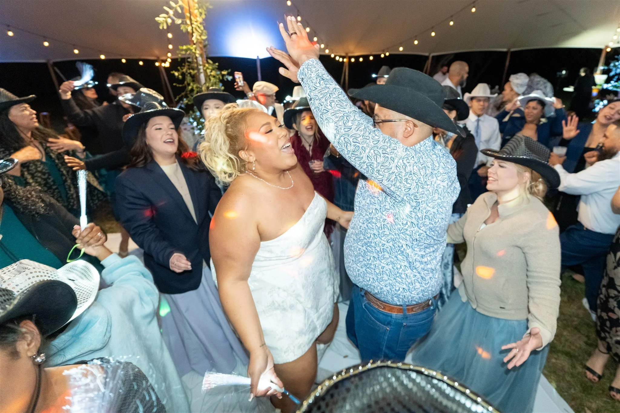 People dancing and celebrating at a party, many wearing cowboy hats, under string lights in a tent.