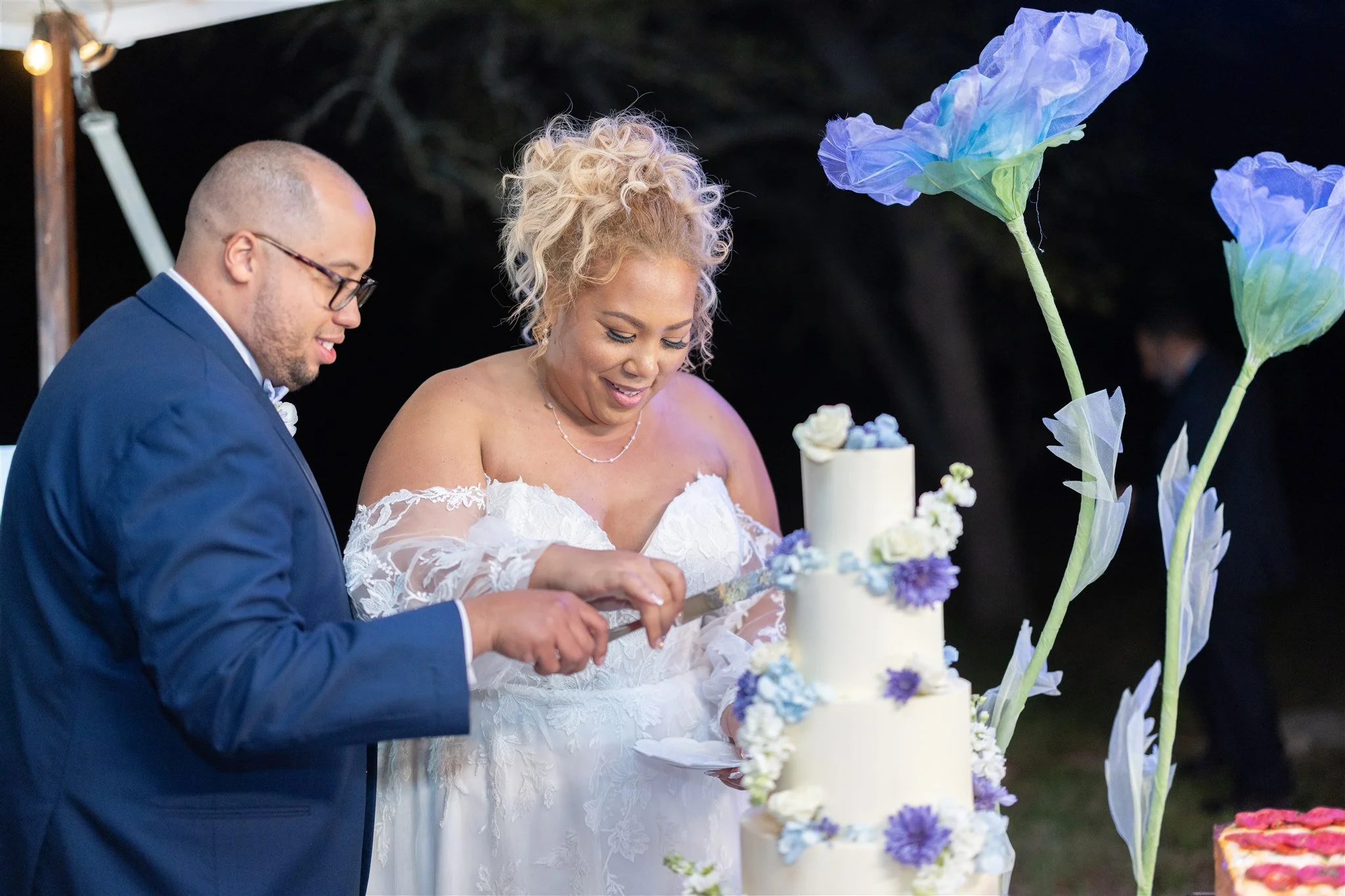 A bride and groom cut a wedding cake outdoors at night, decorated with white and purple flowers, with large artificial blue and purple flowers nearby.