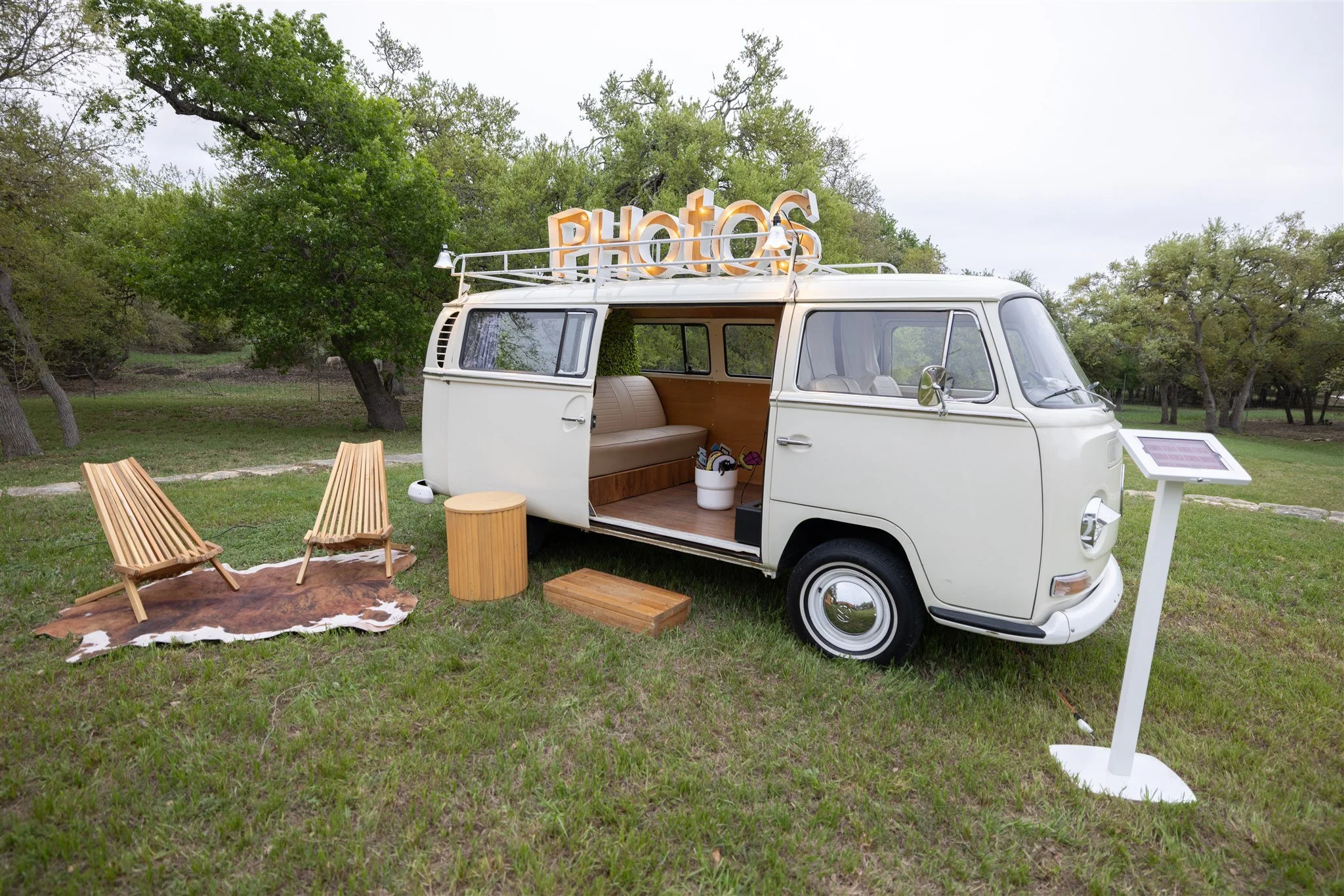 Vintage white Volkswagen camper van with a large sign reading 'PHOTOS' on top, set outdoors on a grassy field with trees in the background. The van's sliding door is open, revealing wood-paneled interior seating. Nearby, there are two wooden lawn cha