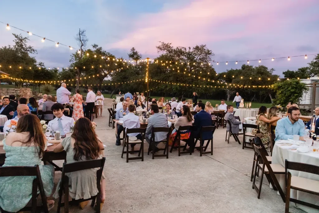 Outdoor evening banquet with round tables, string lights overhead, and guests in semi-formal attire enjoying a meal on a patio surrounded by trees.