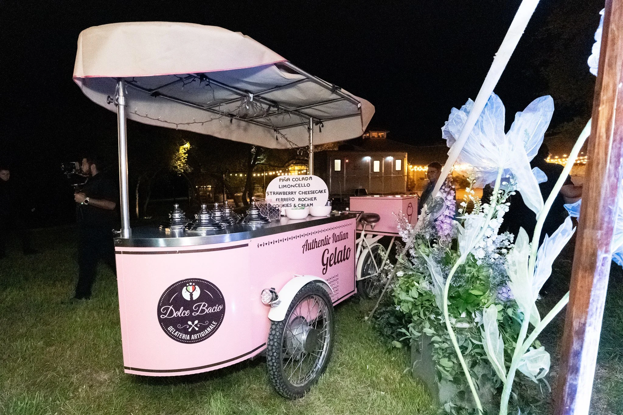 A pink ice cream cart with a white umbrella, labeled 'Dolce Bacio' and 'Authentic Italian Gelato,' is set up outdoors at night. A menu sign lists flavors including Piña Colada, Lemoncello, Strawberry Cheesecake, Ferrero Rocher, and Cookies & Cream. T