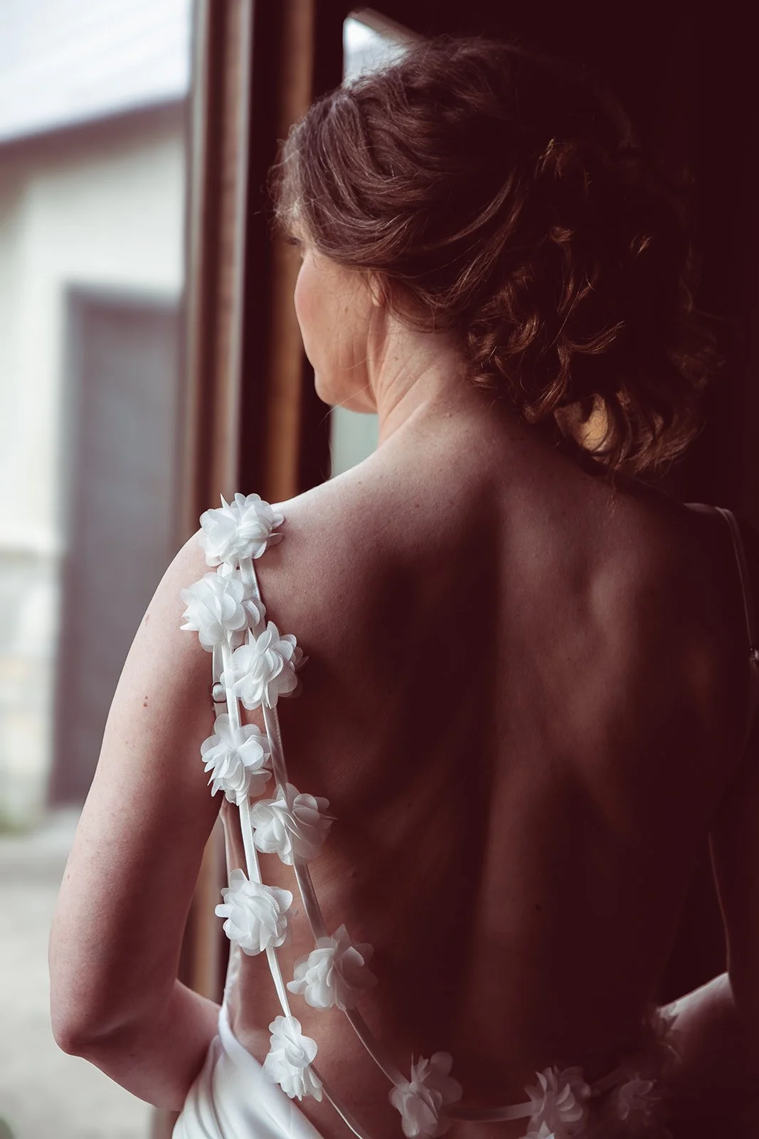 A woman with short, wavy brown hair looking out a window, wearing a backless dress with white floral embellishments on one shoulder.