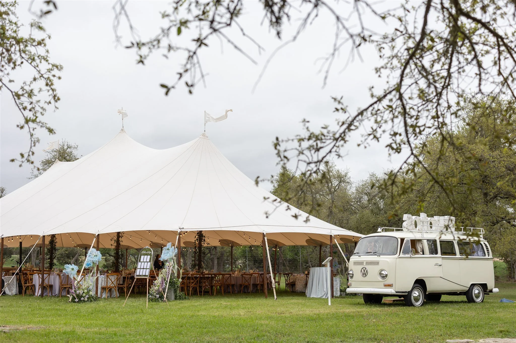 A large white event tent set up outdoors on a grassy area, decorated with floral arrangements and string lights, with a vintage Volkswagen van parked nearby. The scene is outdoors with trees and the sky in the background.