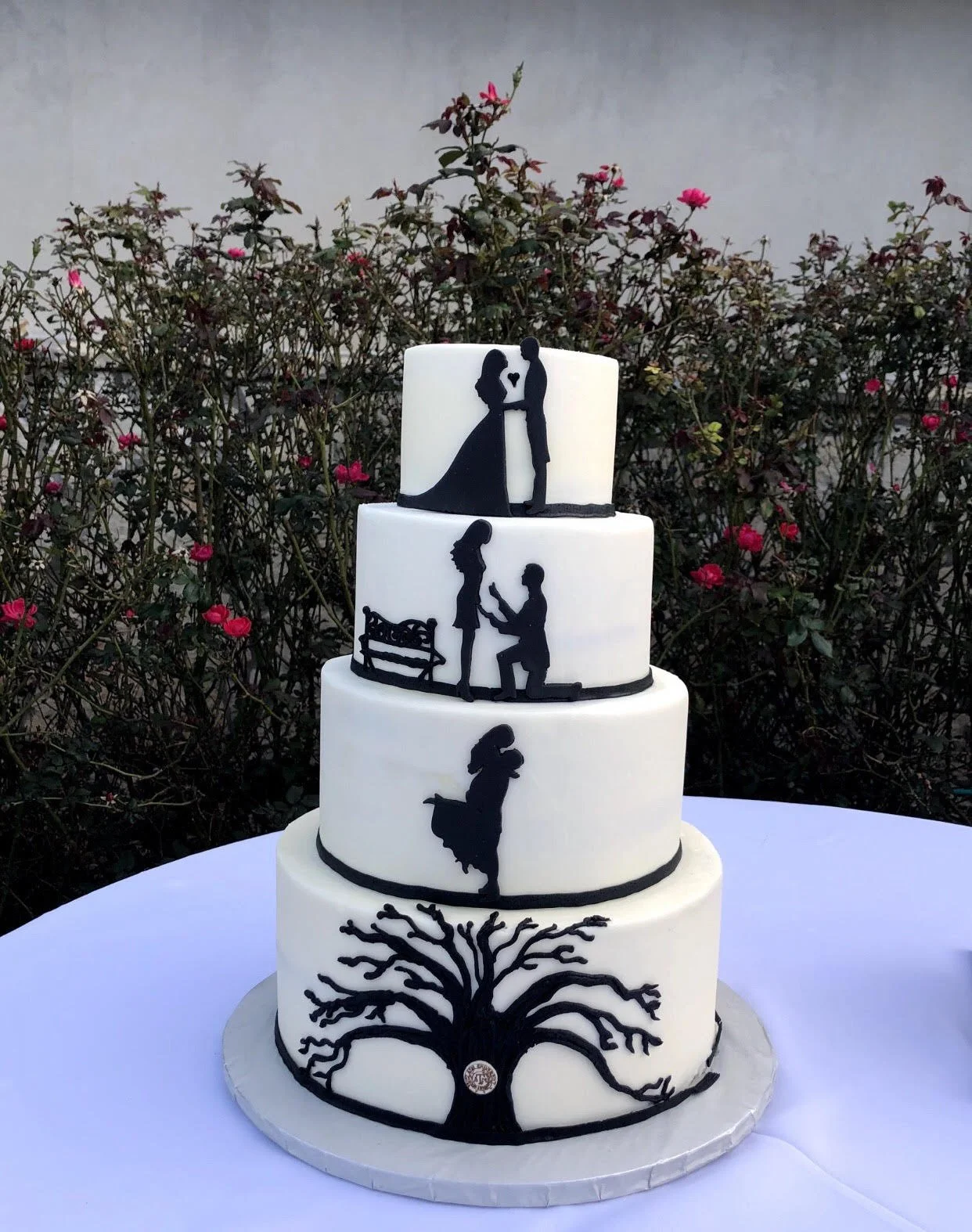 Four-tier white wedding cake decorated with black silhouettes of a mother and daughter, a couple at engagement, a classical dance pose, and a tree at the bottom, set outdoors against a bush with pink flowers.