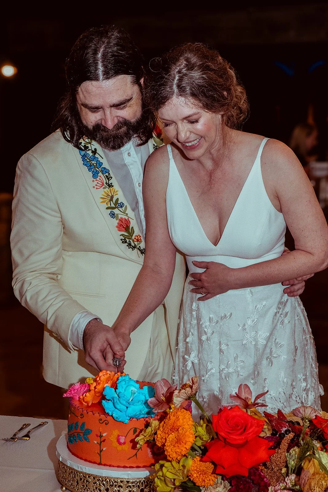 A couple cut a brightly decorated cake together at a celebration, surrounded by flowers.