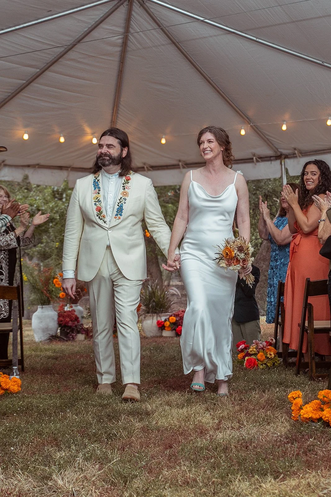Couple walking hand in hand at wedding reception, surrounded by guests clapping and smiling, under a tent decorated with string lights and autumn flowers.