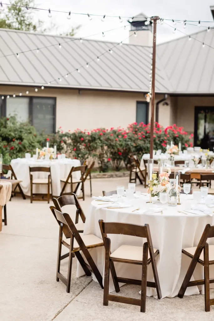 Outdoor event setup with round tables covered in white tablecloths, decorated with flowers and glassware, surrounded by wooden chairs, with string lights overhead and greenery and a building with a metal roof in the background.