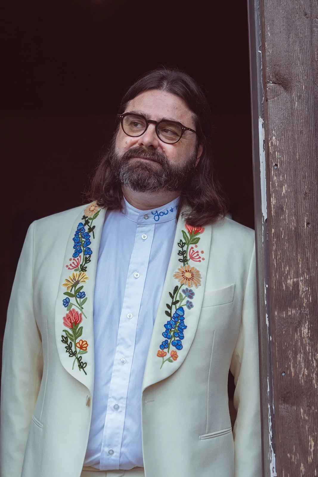 A man with long hair, glasses, and a beard, wearing a white embroidered blazer over a white shirt, standing next to a wooden post.