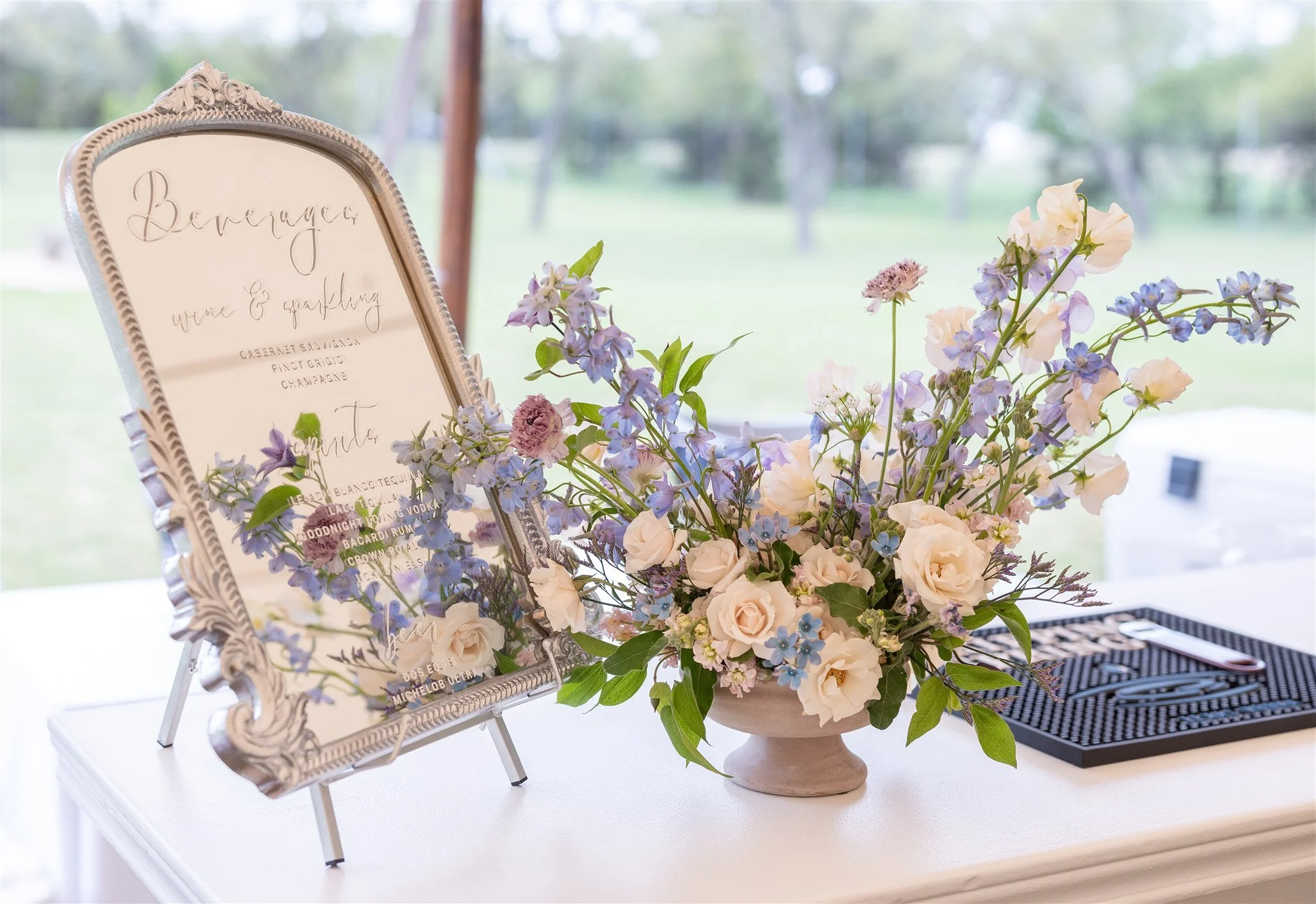 A silver tray with a beverage and quenching sign, a floral arrangement of white roses, light purple and blue flowers, and green leaves, and a black tray with a knife, set on a white table outside with blurred greenery in the background.