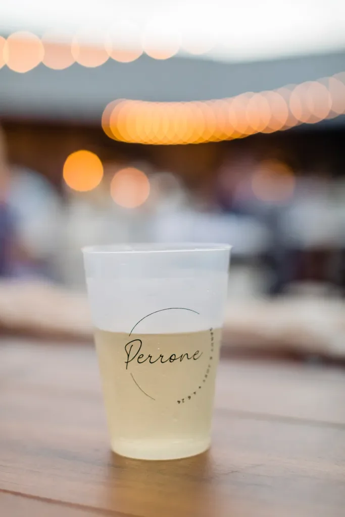 A plastic cup filled with white wine on a wooden table, with the word "Perrone" printed on it, blurred outdoor background with string lights.