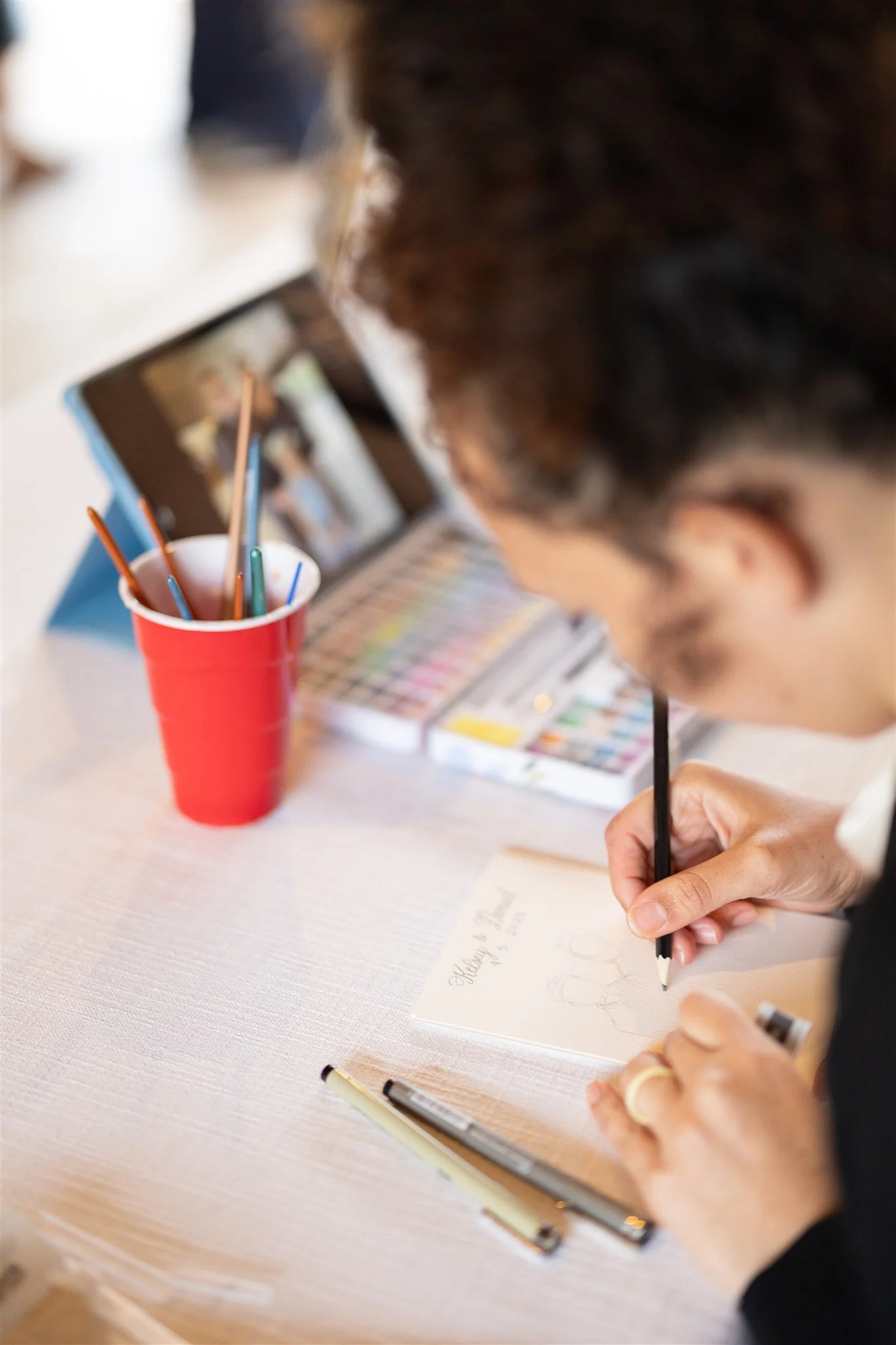 A person is sketching in a notebook with a pen, surrounded by art supplies including colored pencils in a red cup, a set of watercolor paints, and markers on a white table.