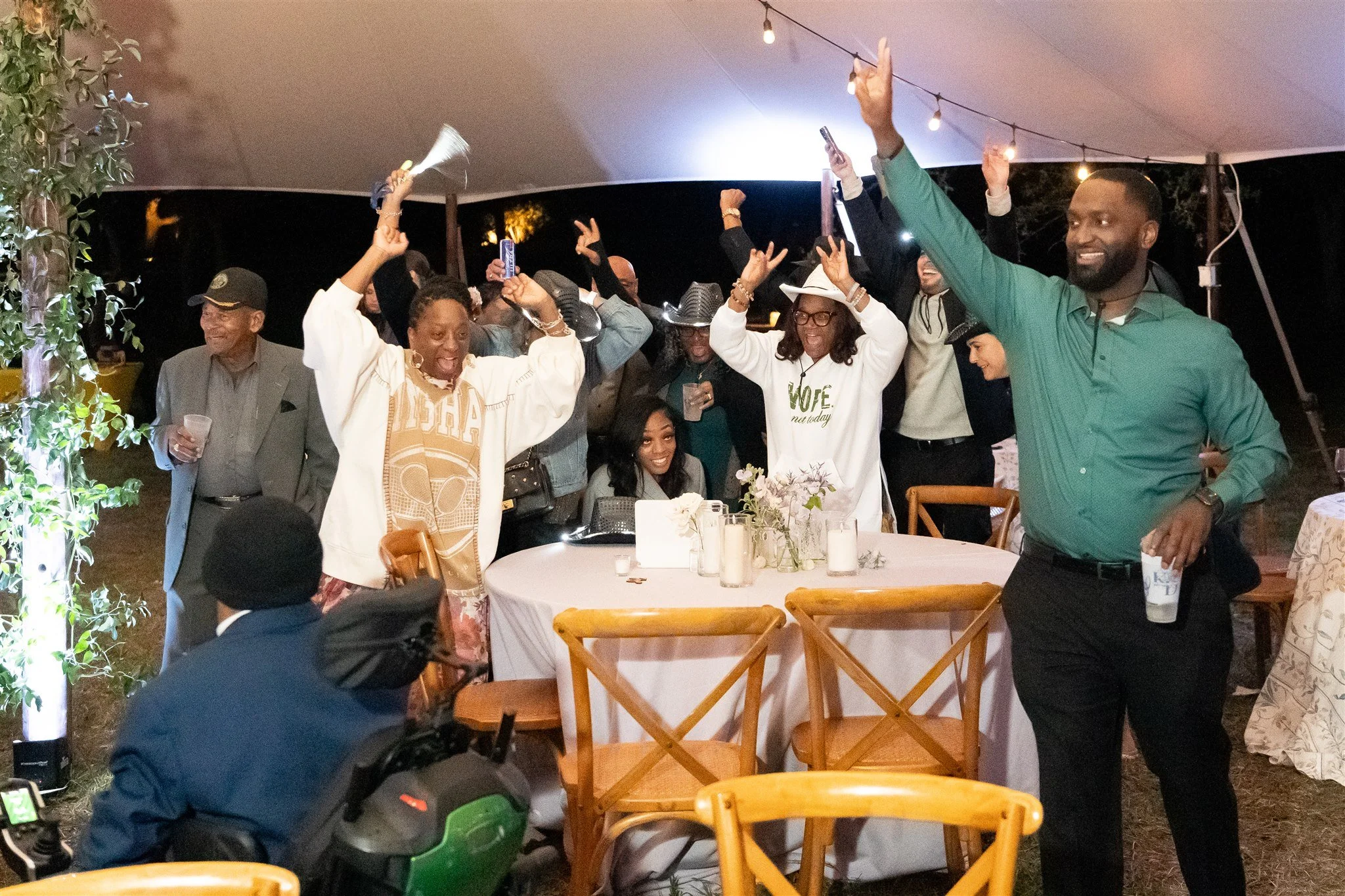 Group of people celebrating around a table at night, some dancing and raising their hands, with a tent and string lights overhead.