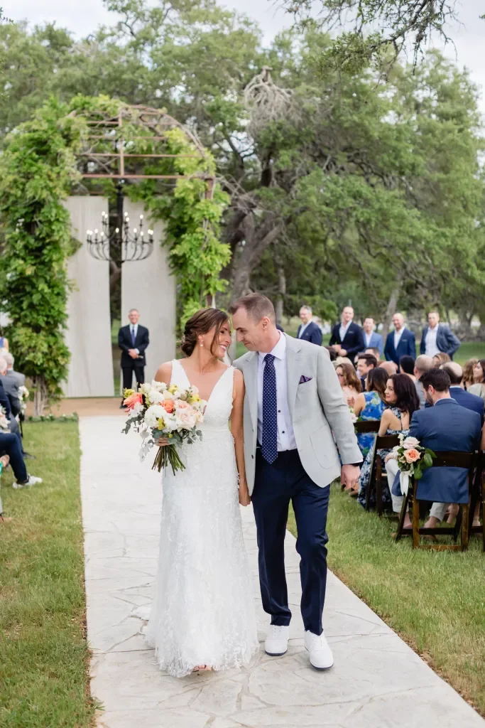 Bride and groom holding hands and smiling during their outdoor wedding ceremony. The bride is in a white lace dress holding a bouquet, and the groom is in a light grey blazer with navy pants and white shoes. Guests are seated on either side, and a fl