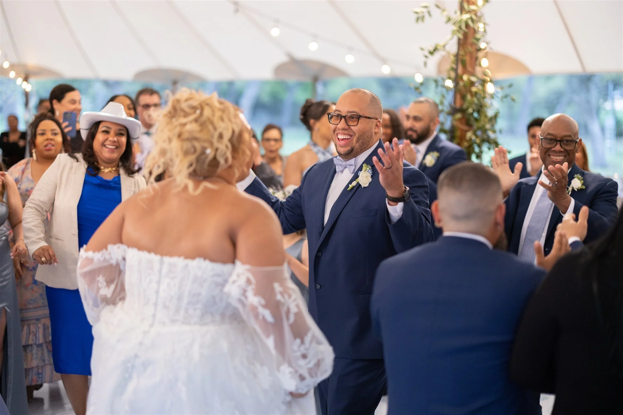 A joyful wedding reception with a group of people dancing and clapping under a large white tent. The focus is on a man in a blue suit and glasses smiling and clapping while facing a woman in a white off-shoulder wedding dress. Guests in the backgroun