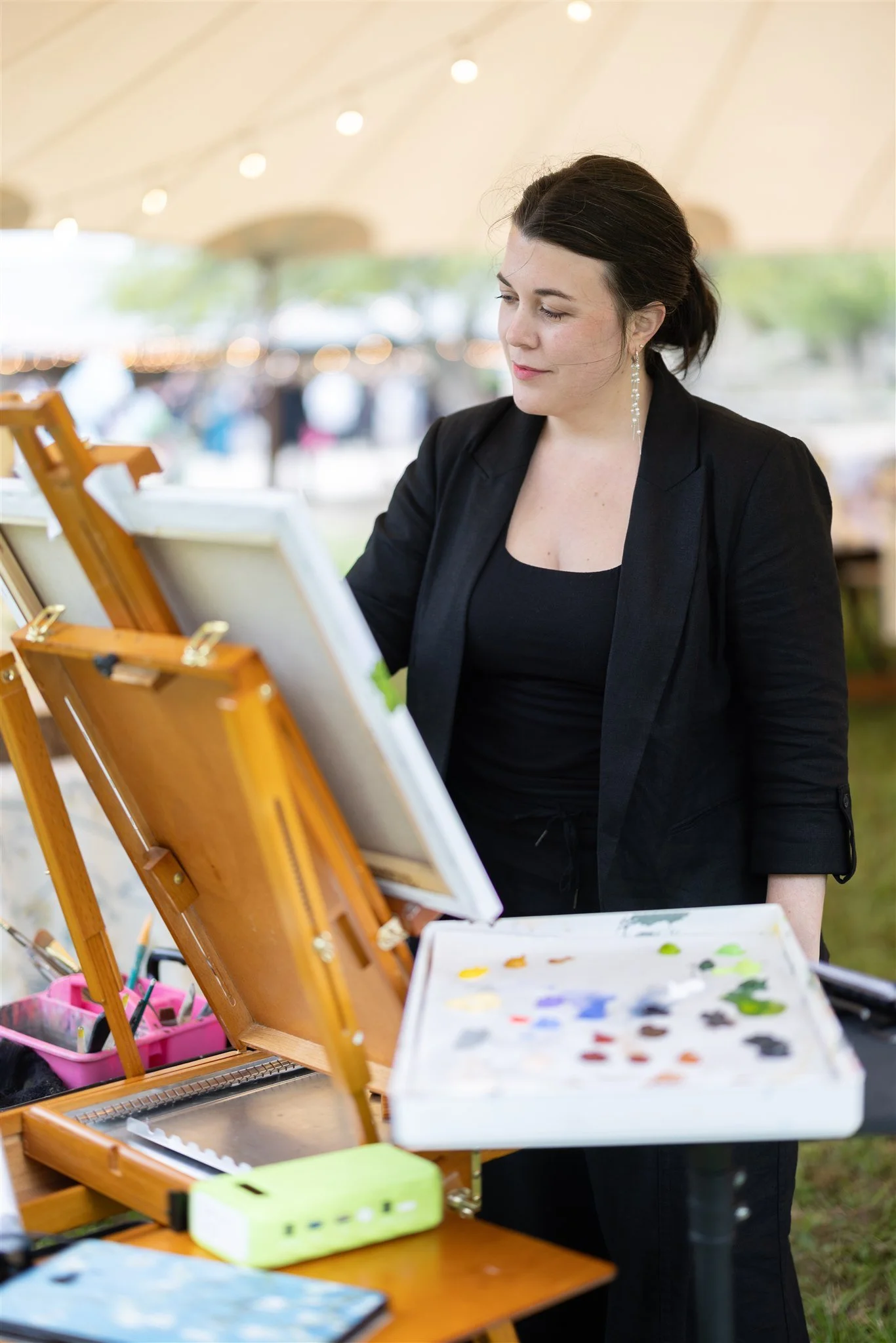 A woman in a black blazer standing outdoors under a canopy, working on a painting at an art station with paints and brushes.