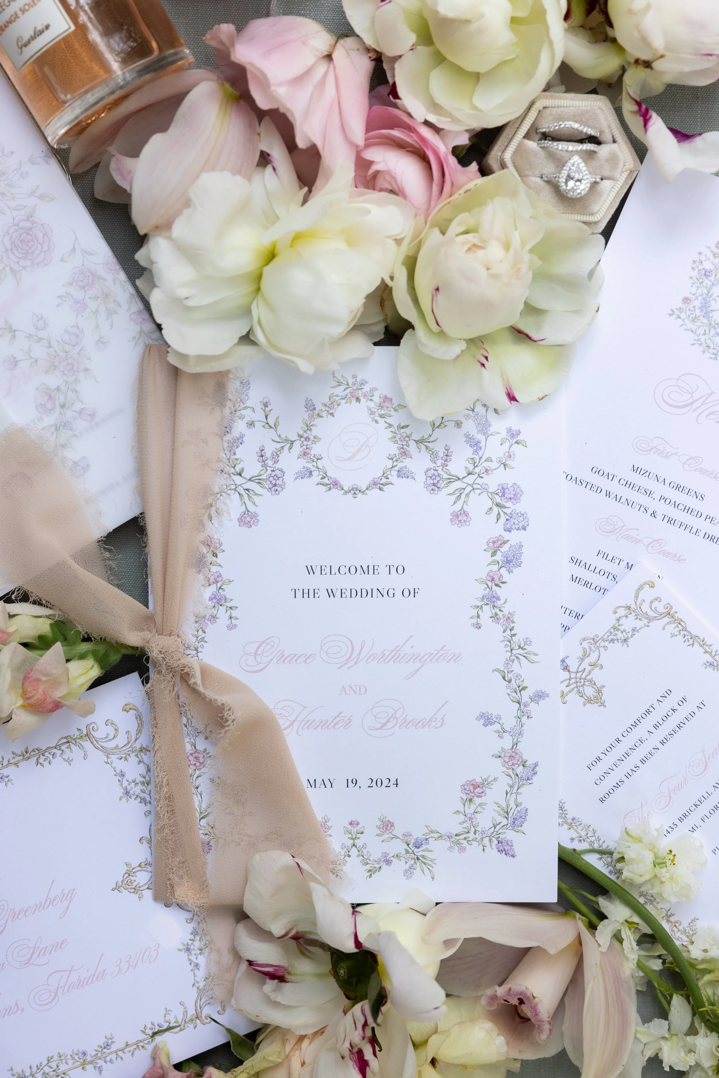 Wedding invitation surrounded by white and pink flowers, with a beige ribbon, and a ring box containing two rings.