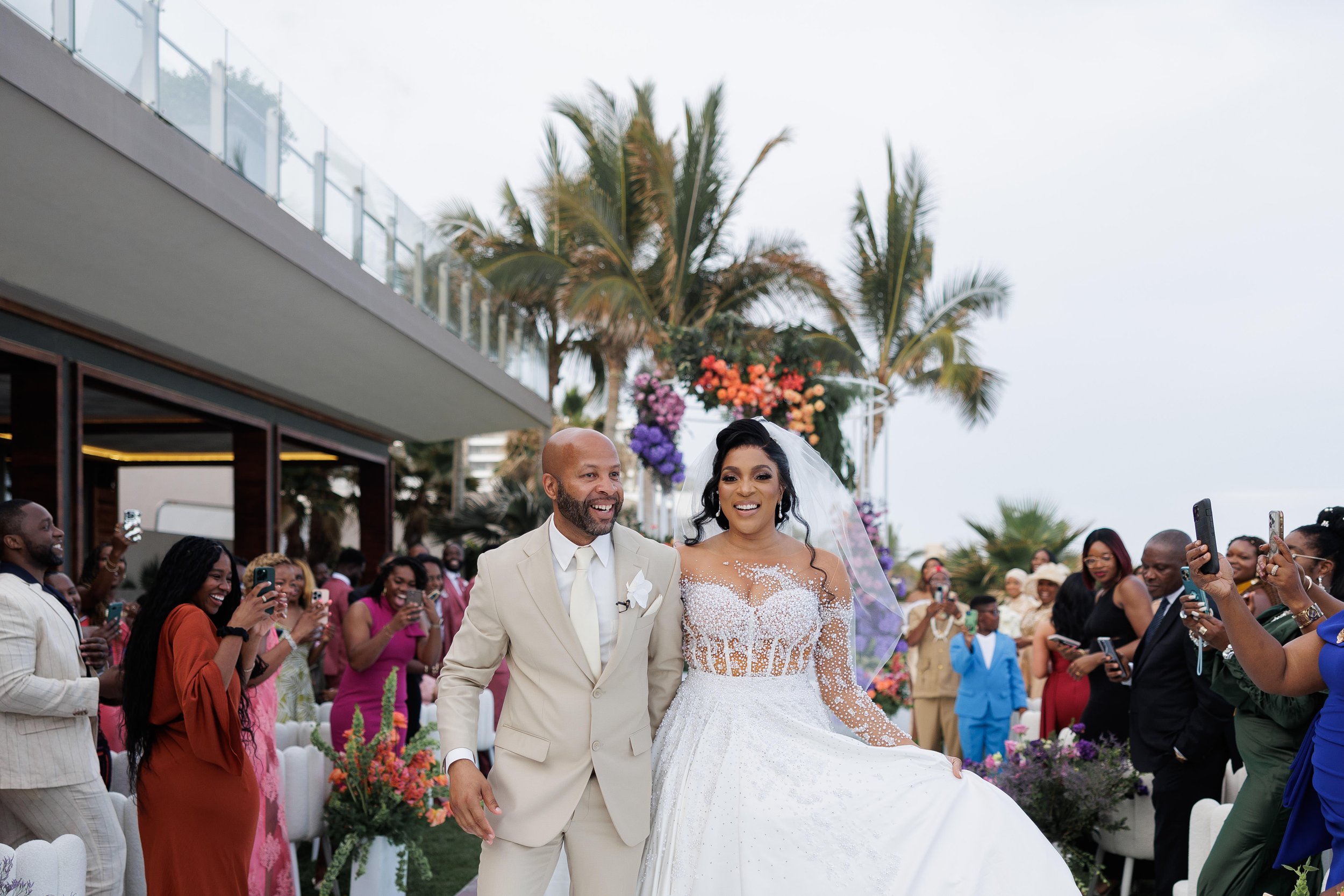 A bride in a white wedding dress and a groom in a beige suit walking down the aisle, smiling, during an outdoor wedding ceremony with guests taking photos and palm trees in background.