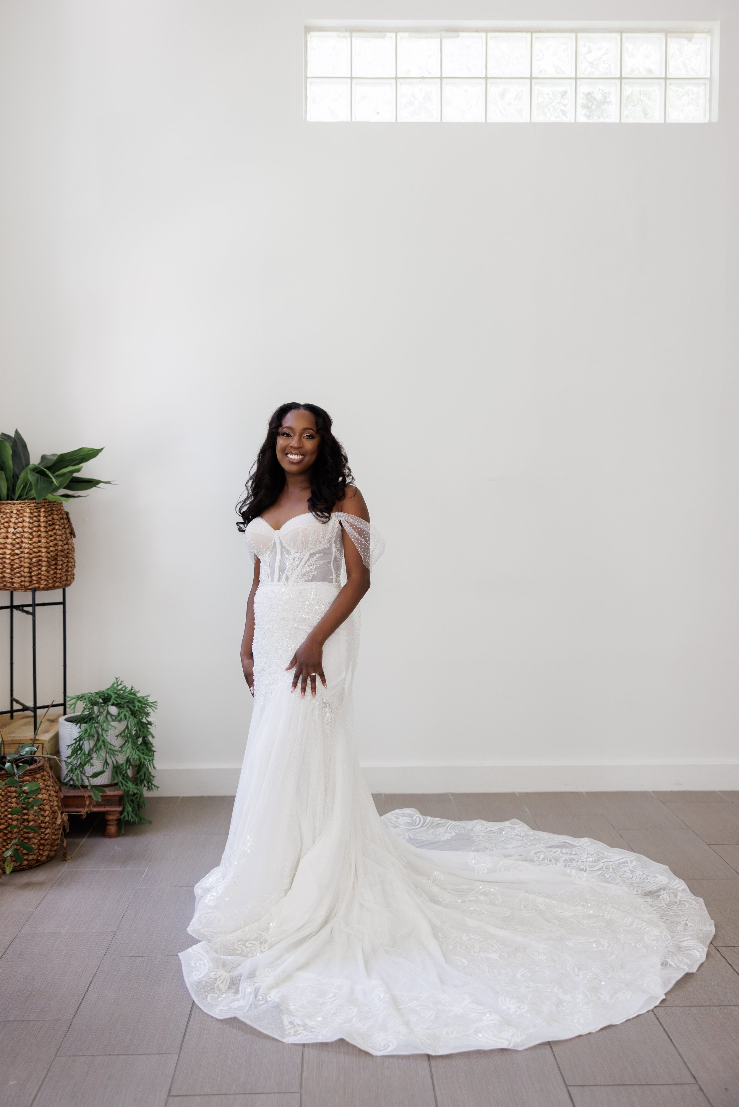 A woman in a white wedding dress standing indoors, smiling, with a plain white wall and a window in the background.