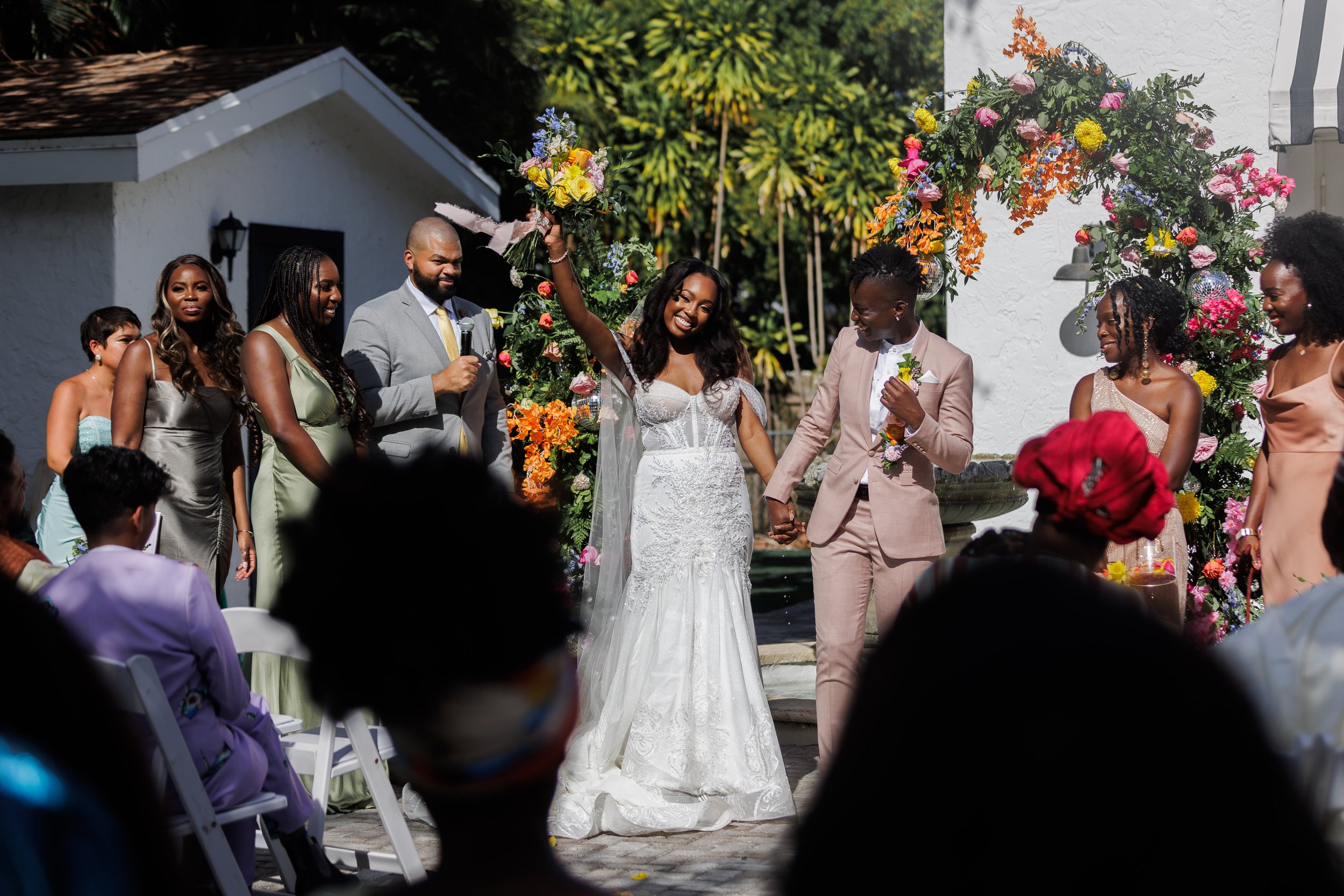 A wedding ceremony outdoors with a bride in a white gown and a groom in a beige suit holding hands, surrounded by friends and family, colorful floral arrangements, and a white building in the background.