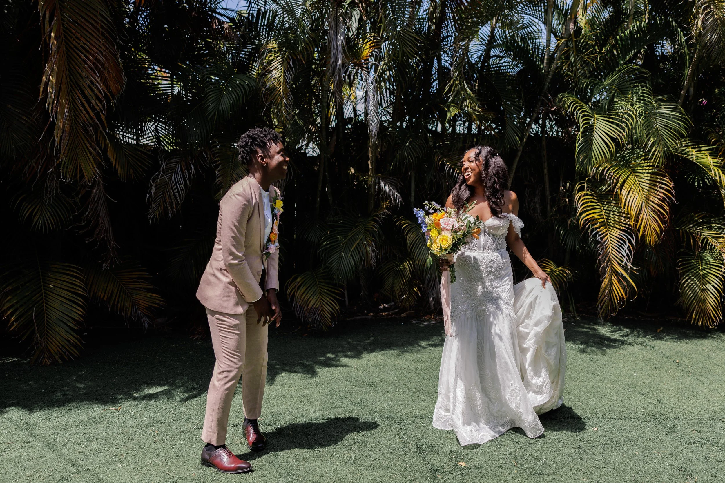 A joyful wedding scene with a bride holding a colorful bouquet and a groom laughing at her in front of lush green tropical plants.