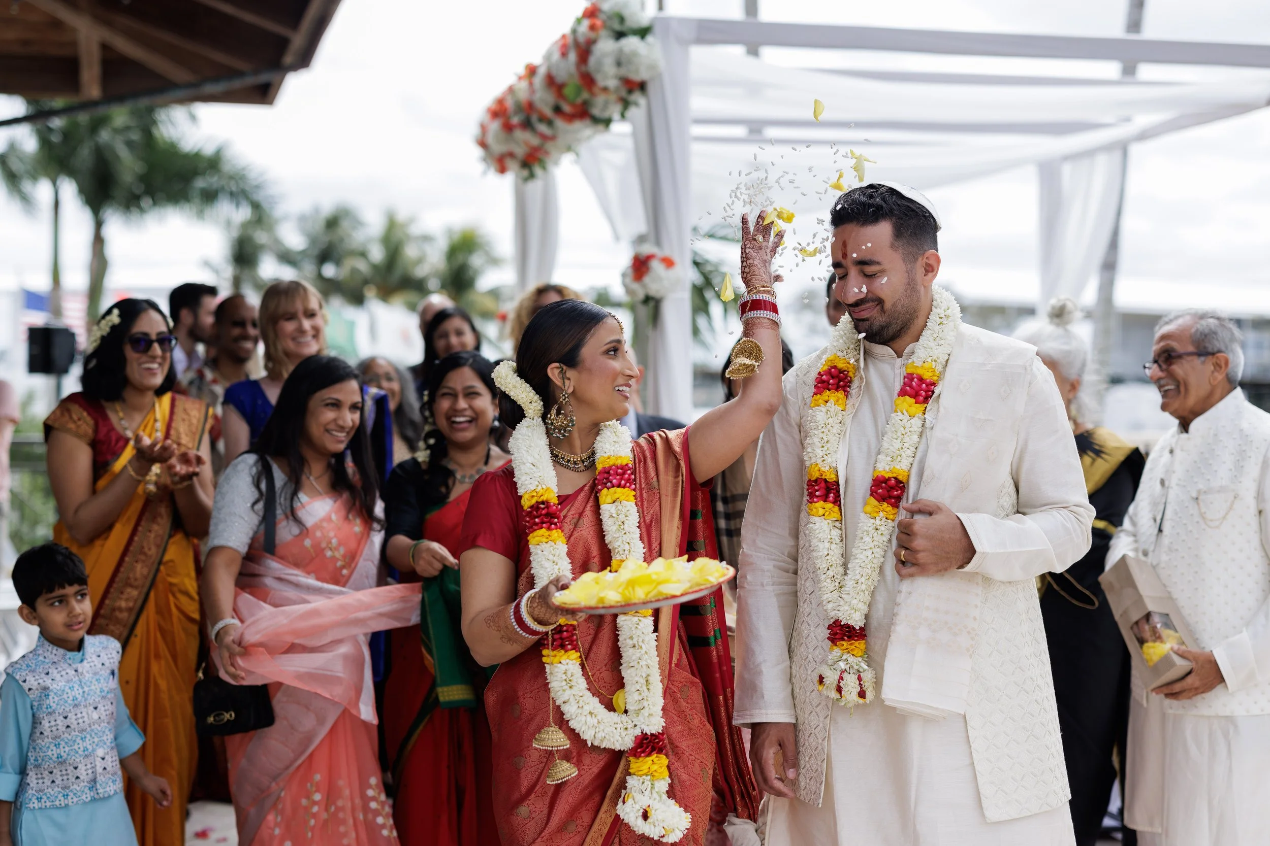 Indian wedding ceremony with bride and groom adorned in traditional attire and garlands, surrounded by family and friends celebrating outdoors.