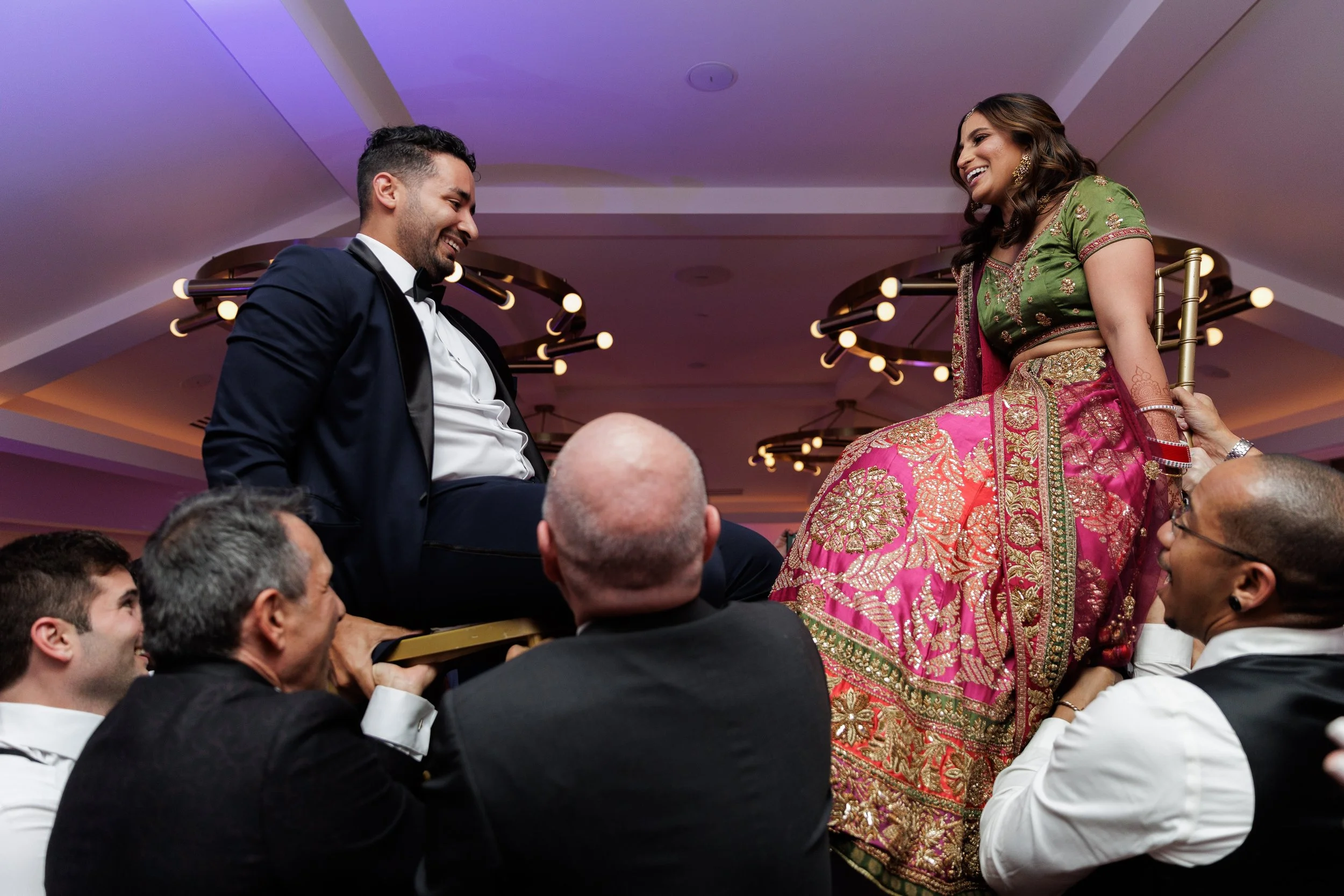 People participating in a traditional Indian celebration, with a man in a tuxedo and a woman in colorful Indian attire seated on a decorated chair being lifted by others.