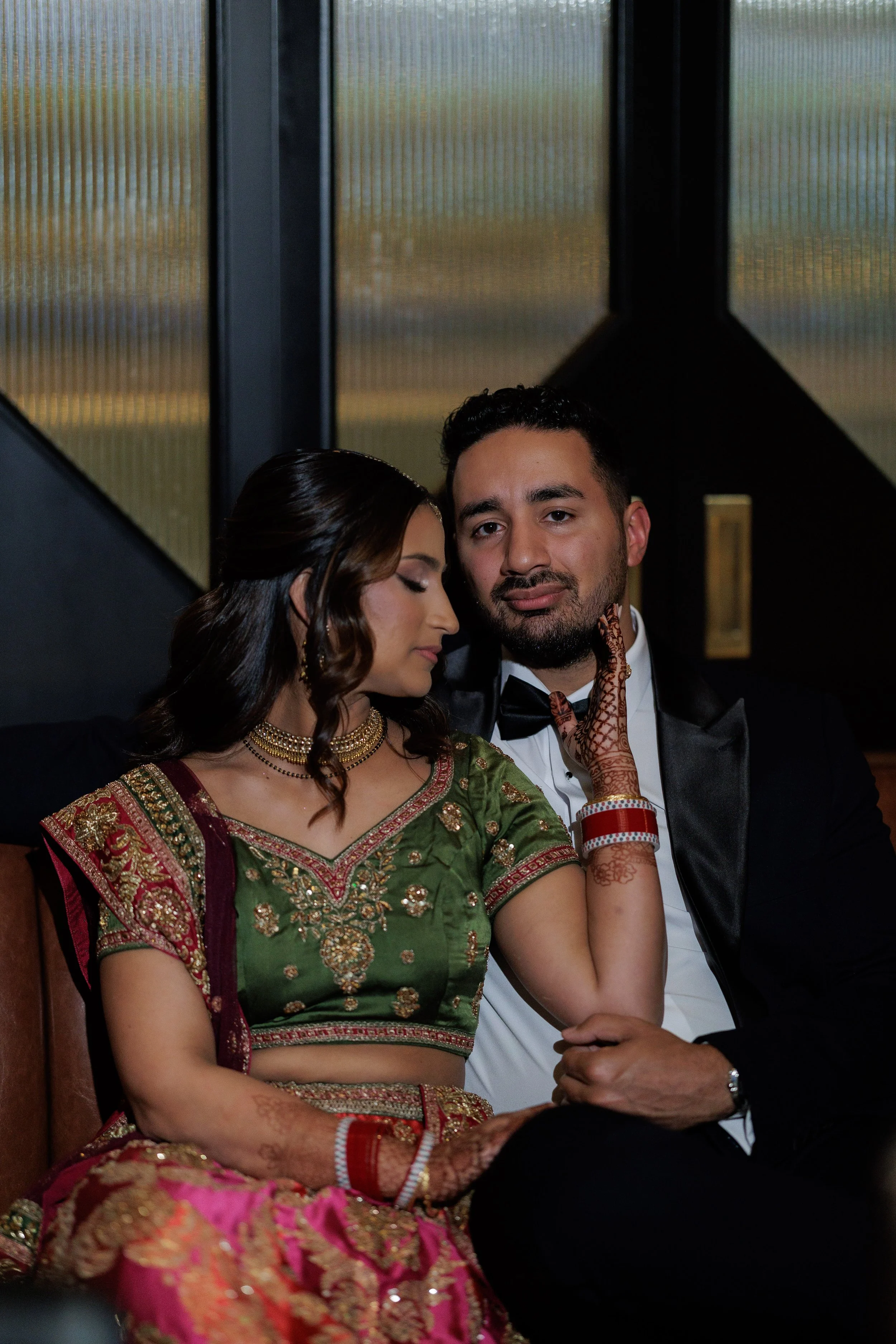 A man and a woman sitting closely together, dressed in formal attire. The woman is wearing traditional Indian clothing with henna on her hands, and the man is in a tuxedo. They appear to be posing for a portrait indoors with large windows in the back