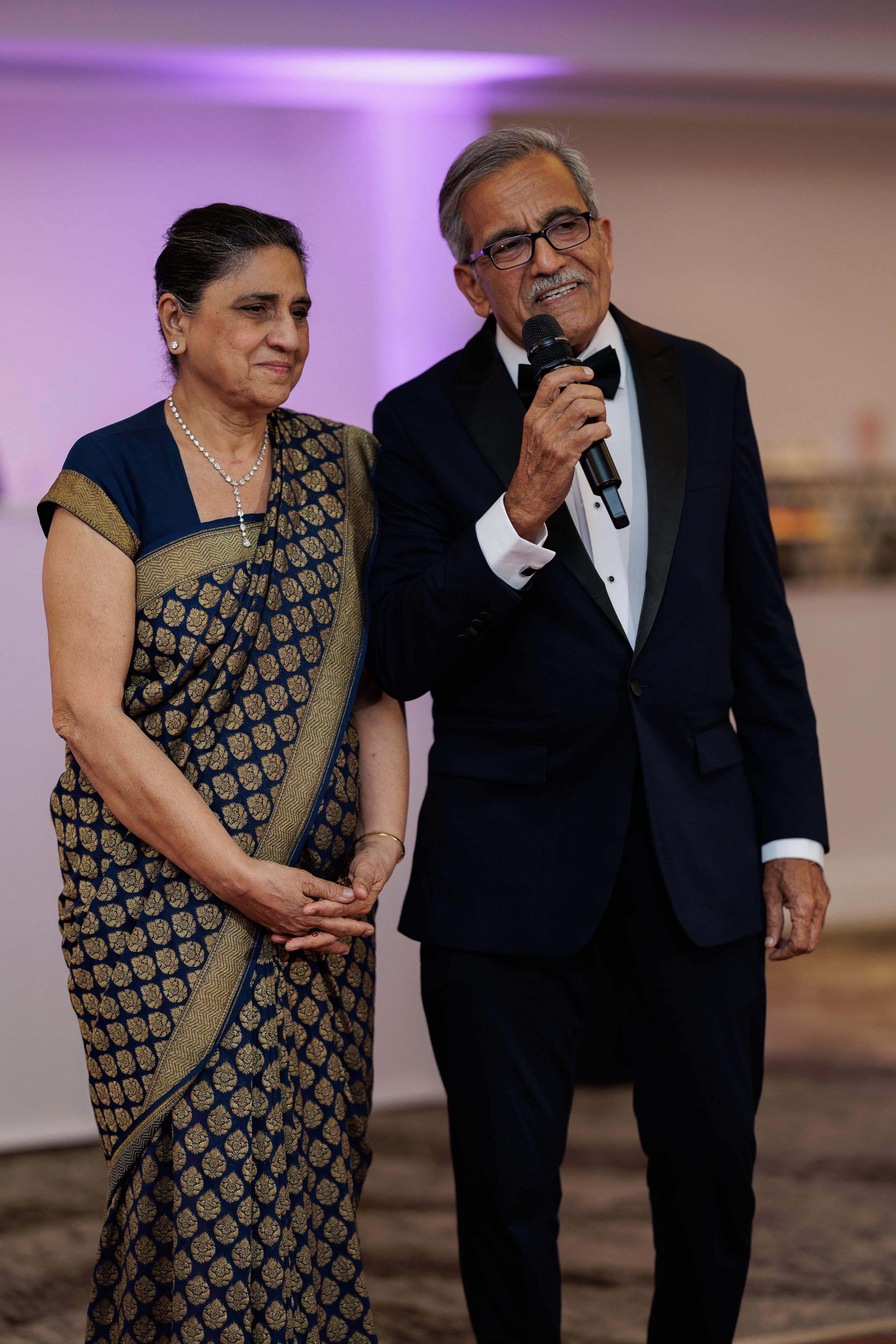 An elderly man in a tuxedo is speaking into a microphone, standing next to an elderly woman in a traditional sari at a formal event.