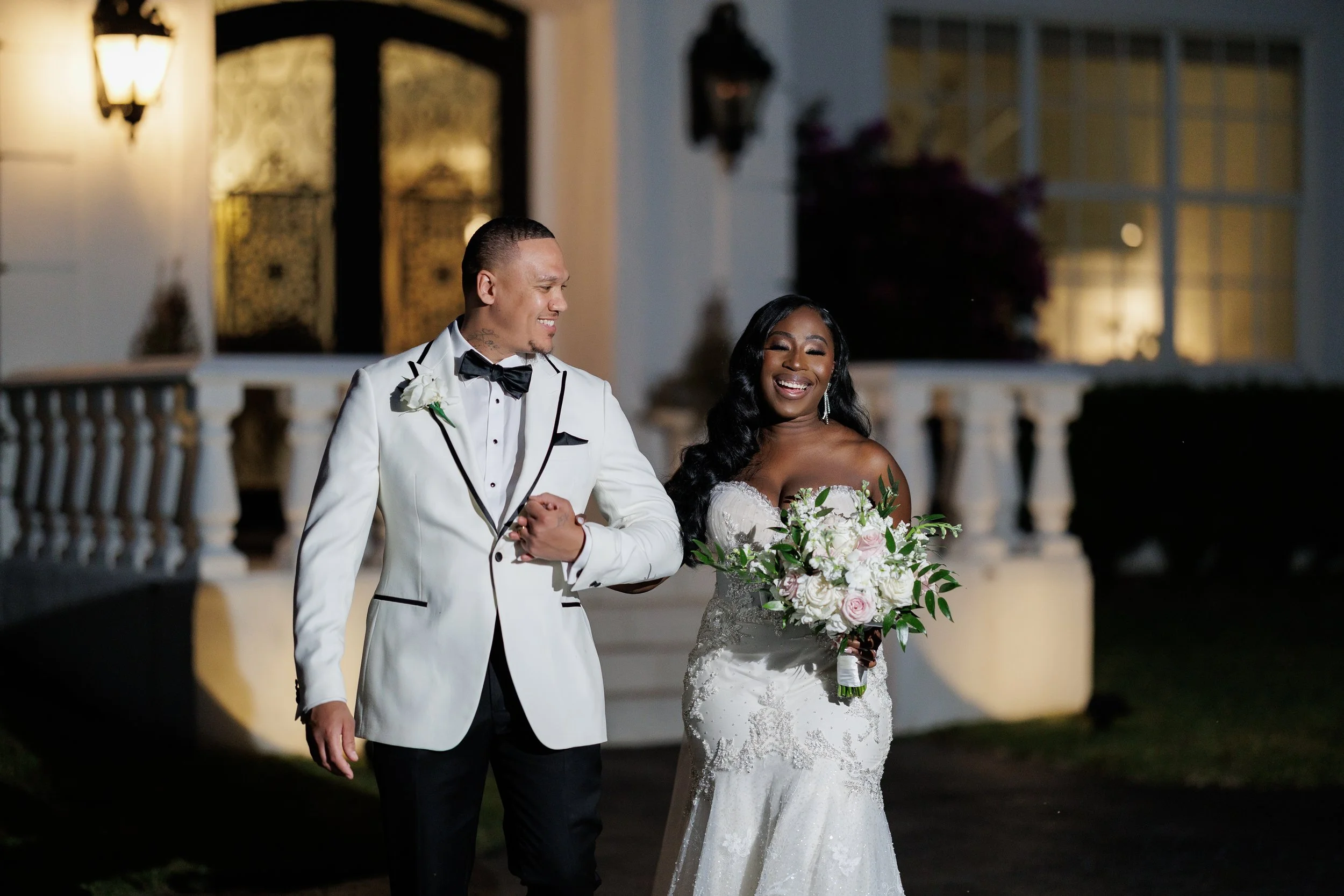 A bride and groom holding hands during their wedding, with the groom kissing the bride's forehead and the bride smiling, surrounded by tropical plants.
