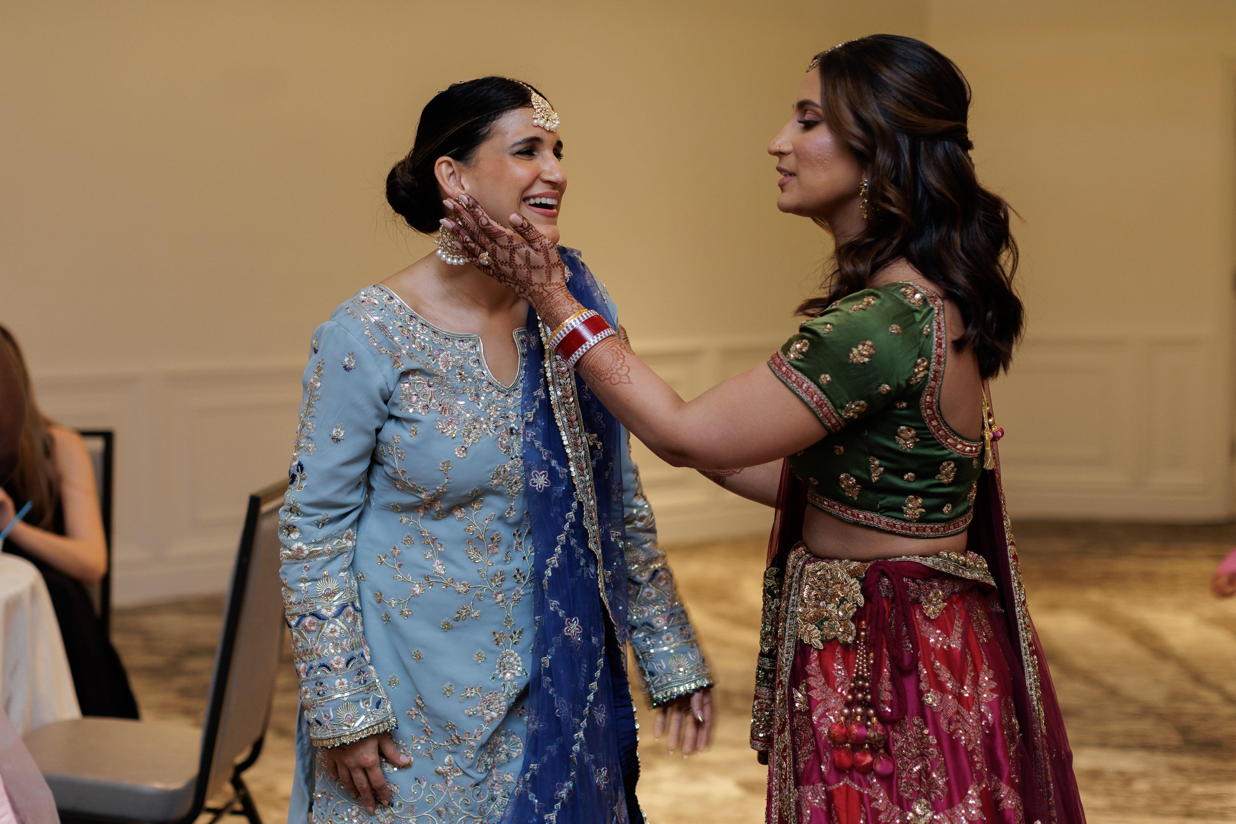 Two women in traditional Indian attire, smiling and embracing at a celebration, with one woman touching the other's face gently.