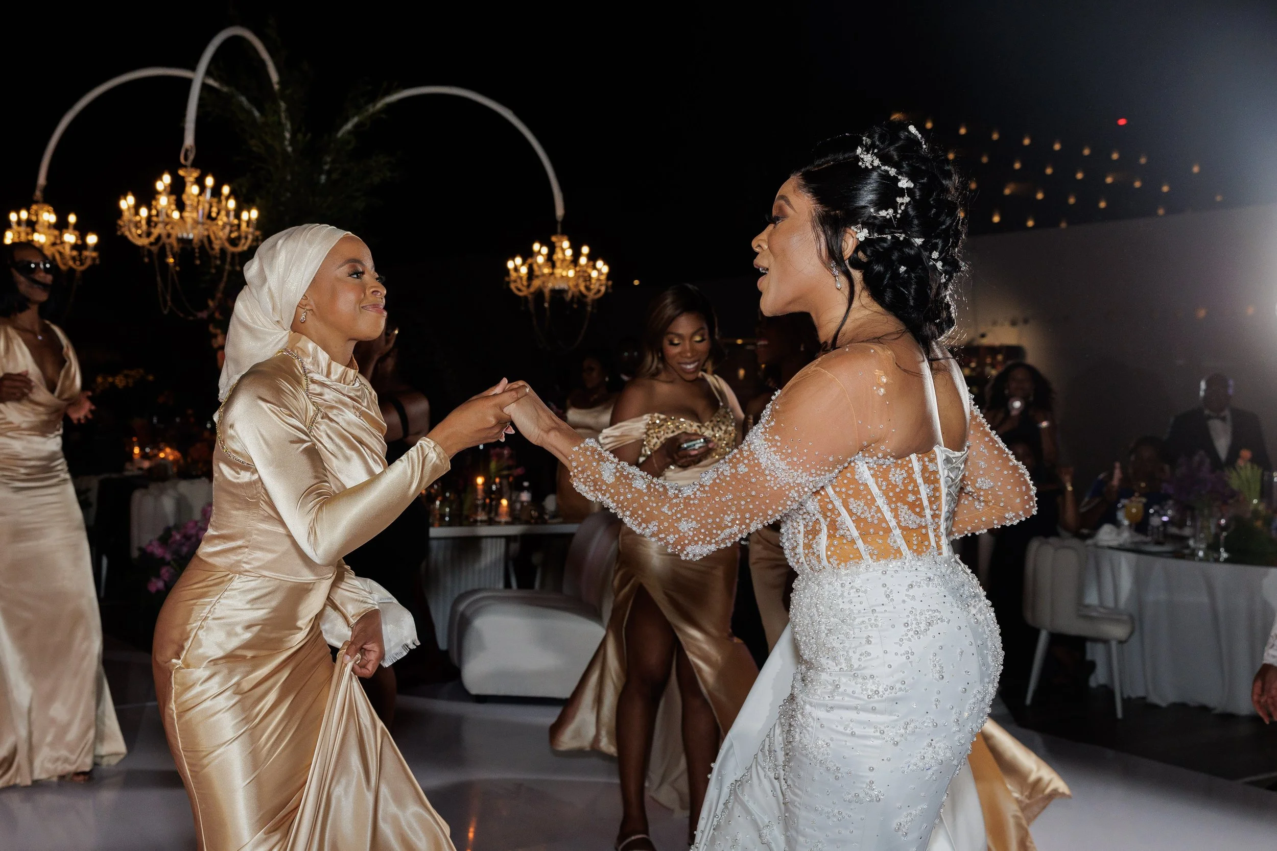 Two women dancing and holding hands at a wedding reception, with other guests in the background, elegant chandeliers hanging from the ceiling, and a nighttime cityscape visible outside.