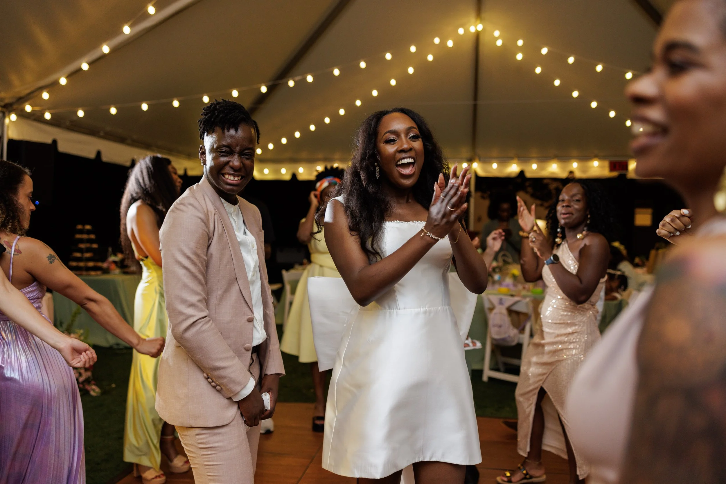 People dancing and celebrating at a wedding reception under string lights in a decorated tent.
