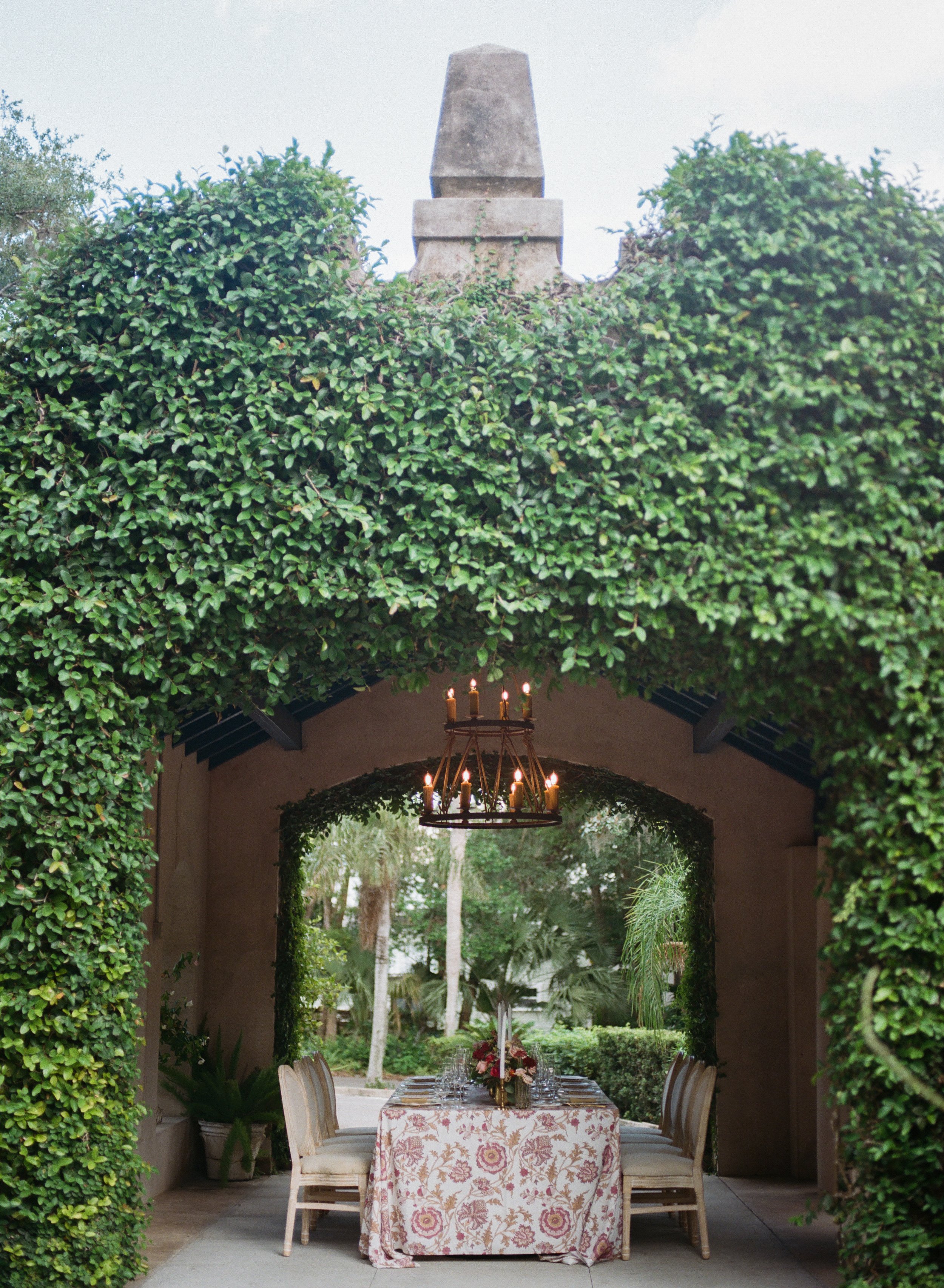 An outdoor dining area under an archway covered with greenery, with a chandelier and seating around a table set with flowers and candles.