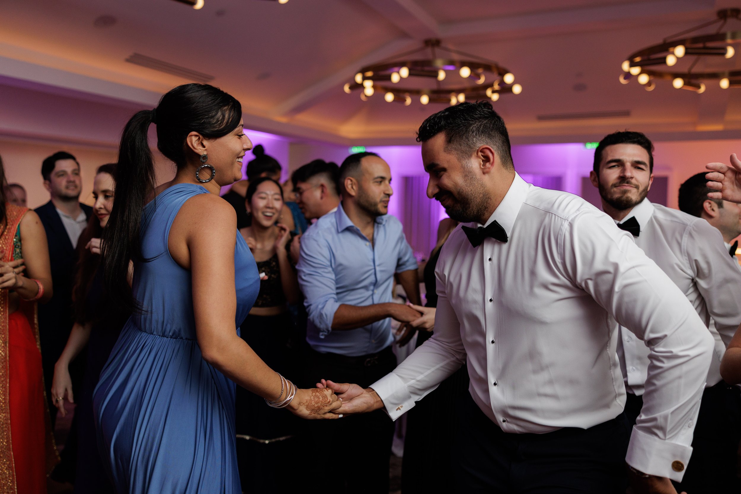 People dancing and celebrating at a wedding reception, with a woman in a blue dress and a man in a white tuxedo shaking hands and smiling.