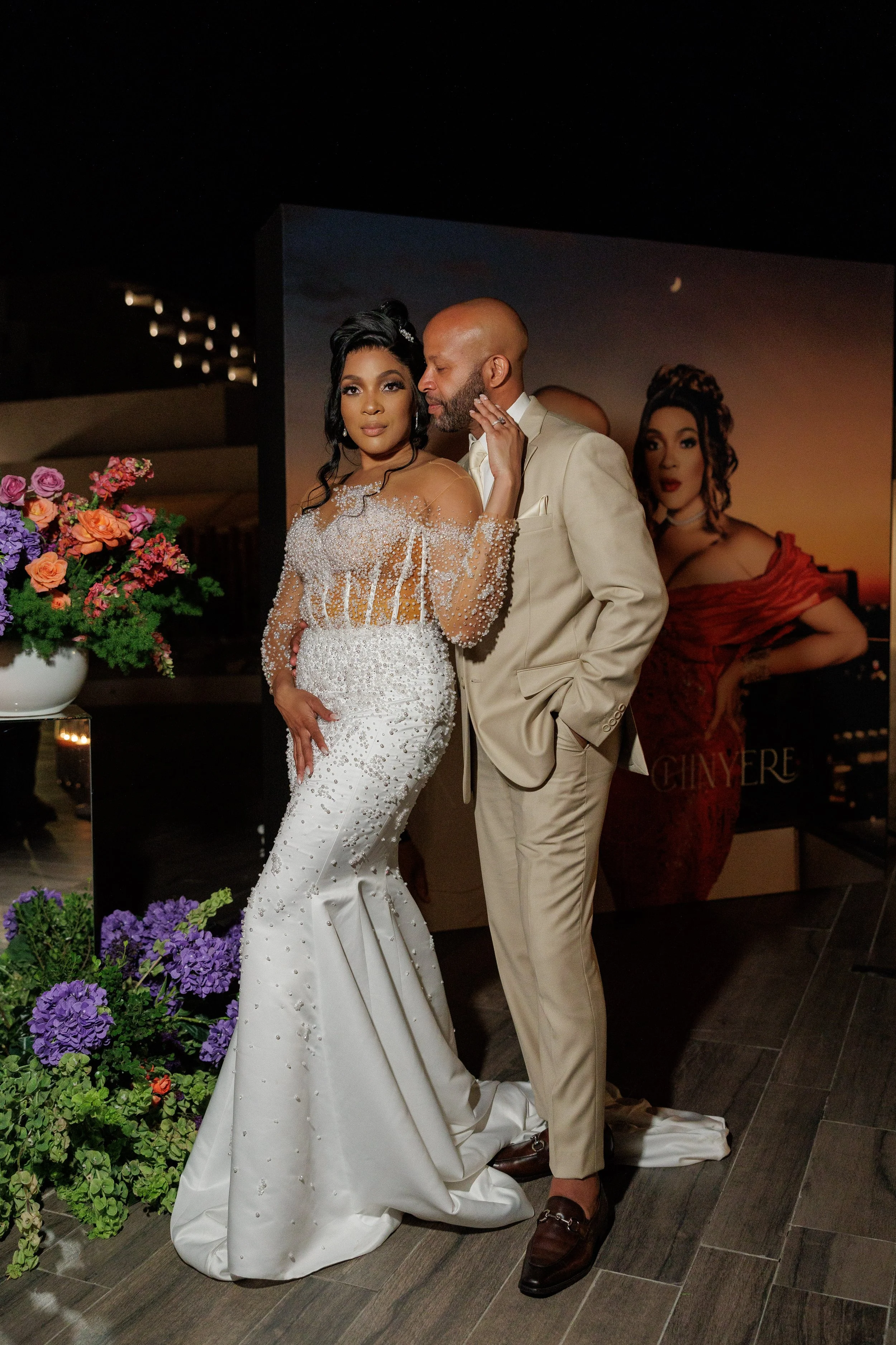 A couple dressed in formal wedding attire poses together indoors. The woman wears a white, intricately beaded wedding gown, and the man is in a beige tuxedo with brown shoes. There are flowers and a large poster of a woman in a red dress in the background.