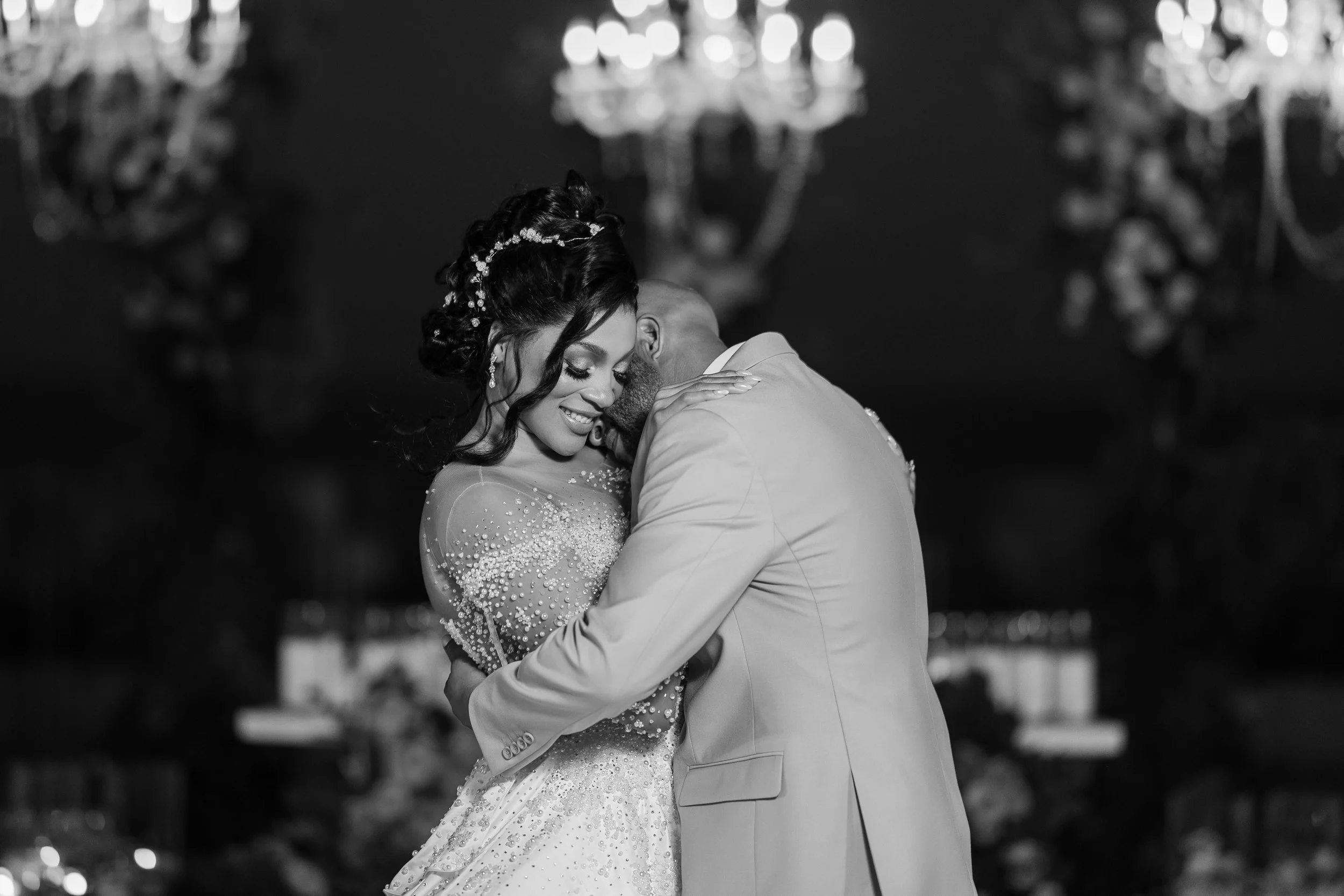 A black and white photo of a bride and groom sharing a dance, close together, smiling with their eyes closed, with chandeliers hanging overhead.
