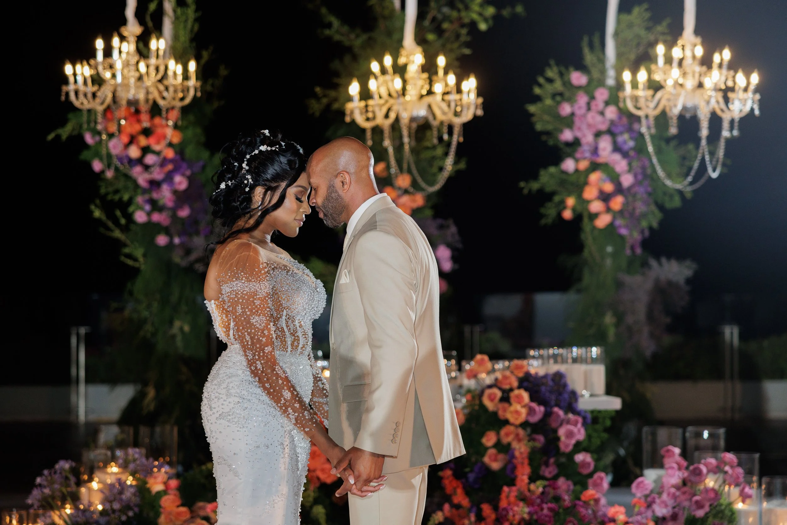 A bride and groom standing closely with their foreheads touching, holding hands during their wedding ceremony at night. The bride is wearing a beaded white gown with sheer sleeves, and the groom is dressed in a cream-colored suit. The background feat