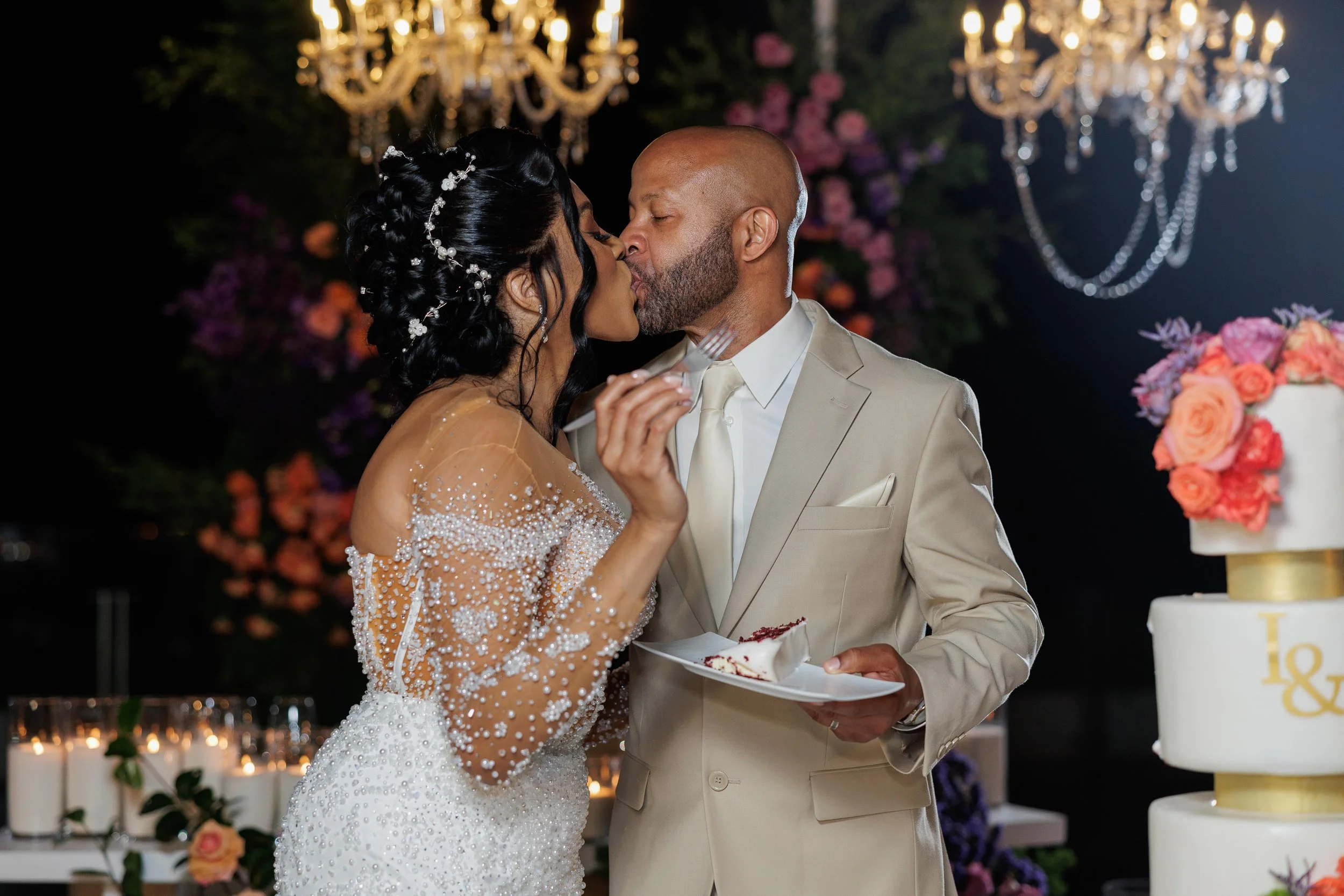 A bride and groom share a kiss at their wedding reception, with a wedding cake and floral decorations in the background.